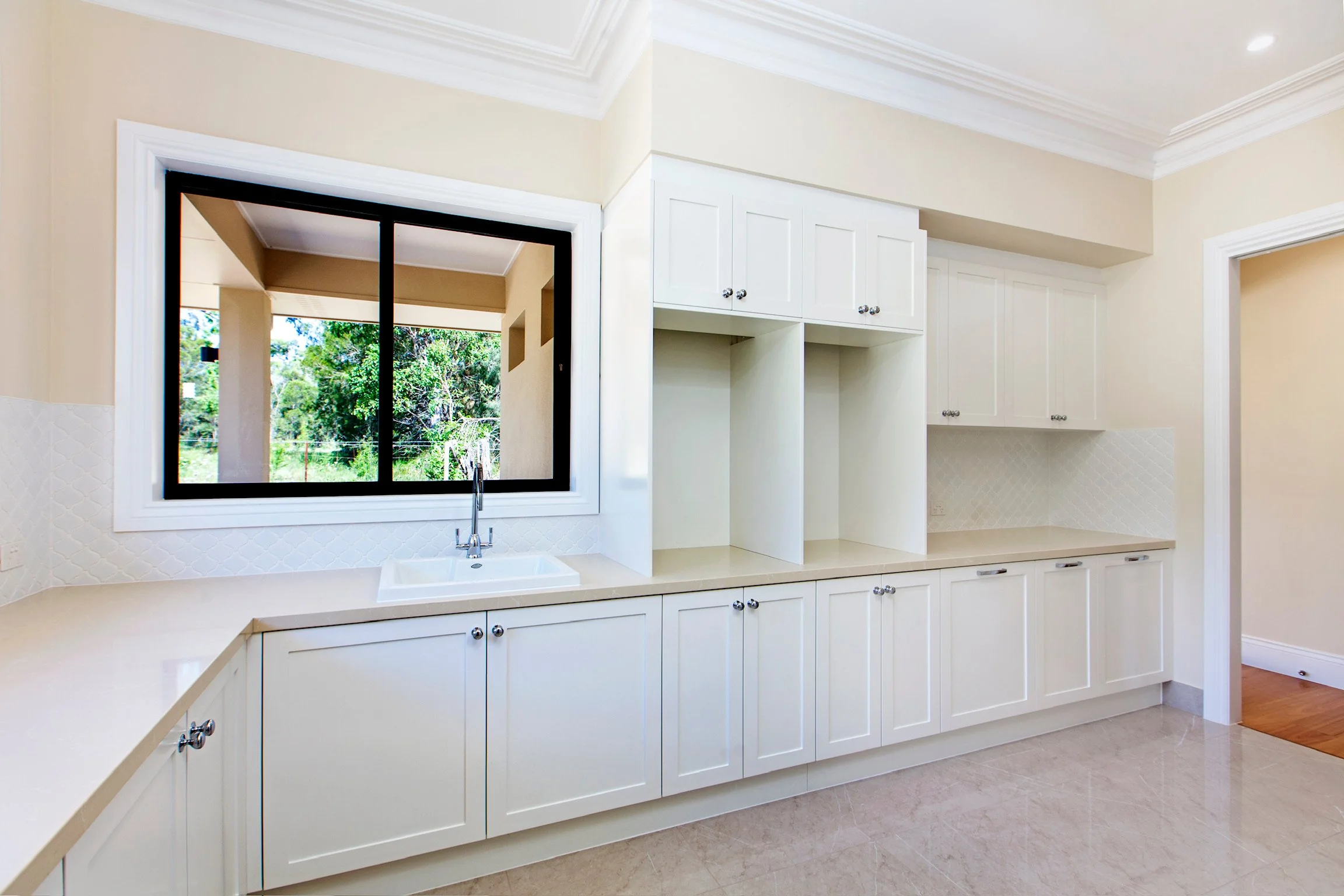A bright kitchen with white cabinets, a window above the sink looking out to greenery, beige countertops, and a light-colored tiled floor.
