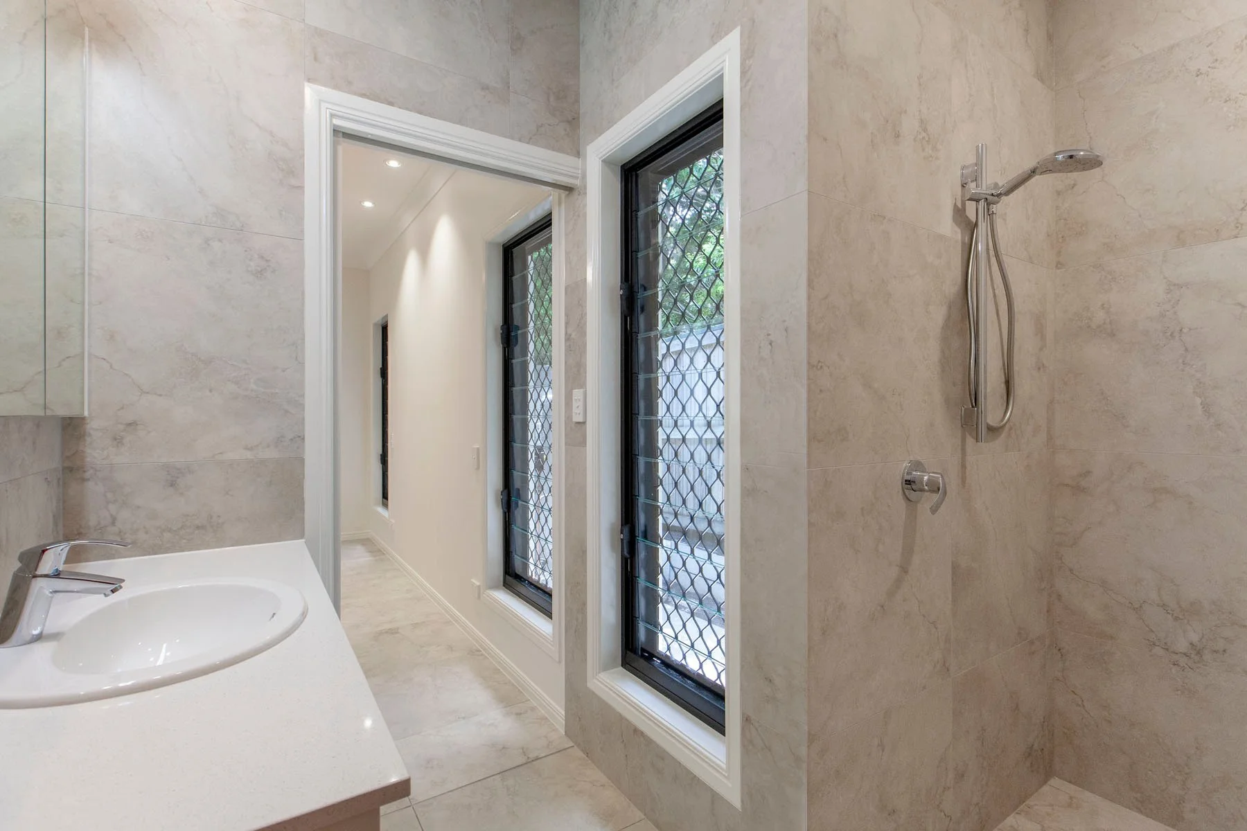 A bathroom with beige marble walls and floor, a white sink with a silver faucet, a window with security bars, and a shower area with a silver showerhead and controls.