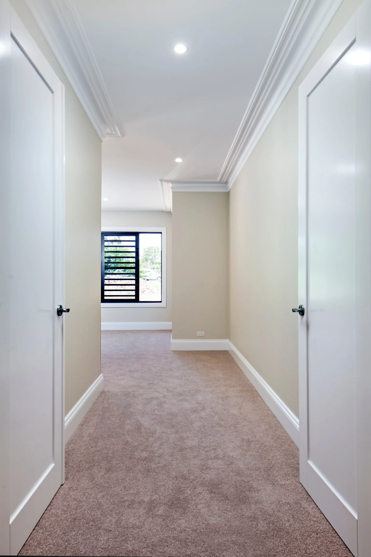 A hallway with beige carpeting, cream-colored walls, crown molding, and two open white doors on either side. At the end of the hallway, there is a window with black horizontal bars, showing outdoor greenery.