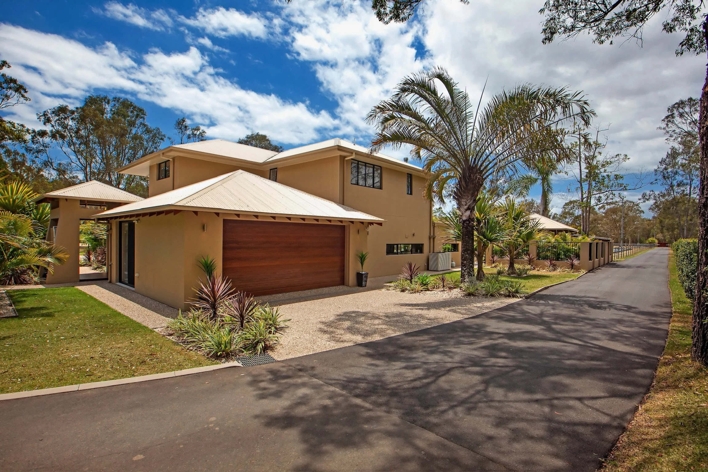 A modern two-story house with beige walls and a brown garage door, surrounded by desert plants and trees, under a partly cloudy blue sky.