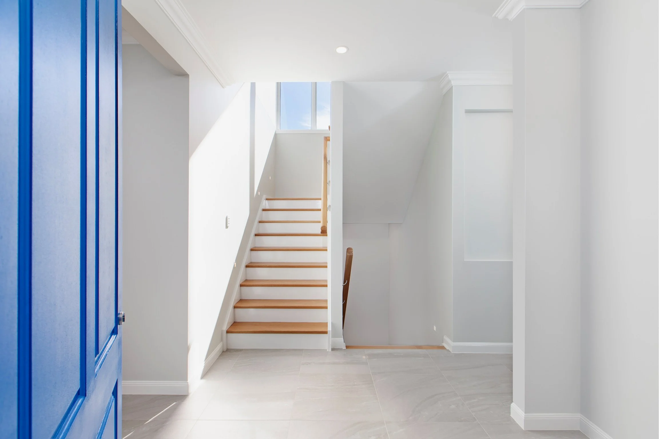 Interior view of a bright, white staircase area with wooden steps, high windows letting in natural light, and a round ceiling light.
