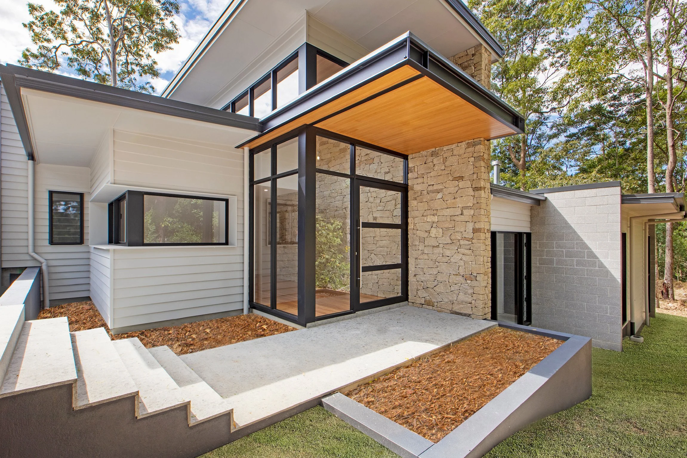 Modern house exterior with stone, white siding, and large glass windows and door, surrounded by trees and outdoor concrete steps.