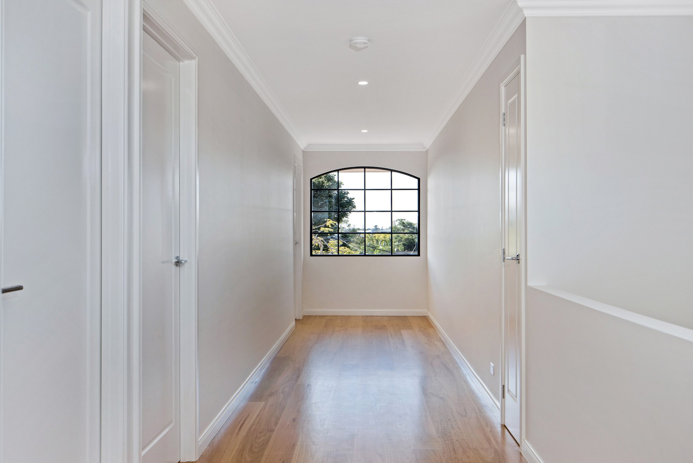 Bright hallway with white walls, hardwood floors, and a large arched window at the end showing trees outside.