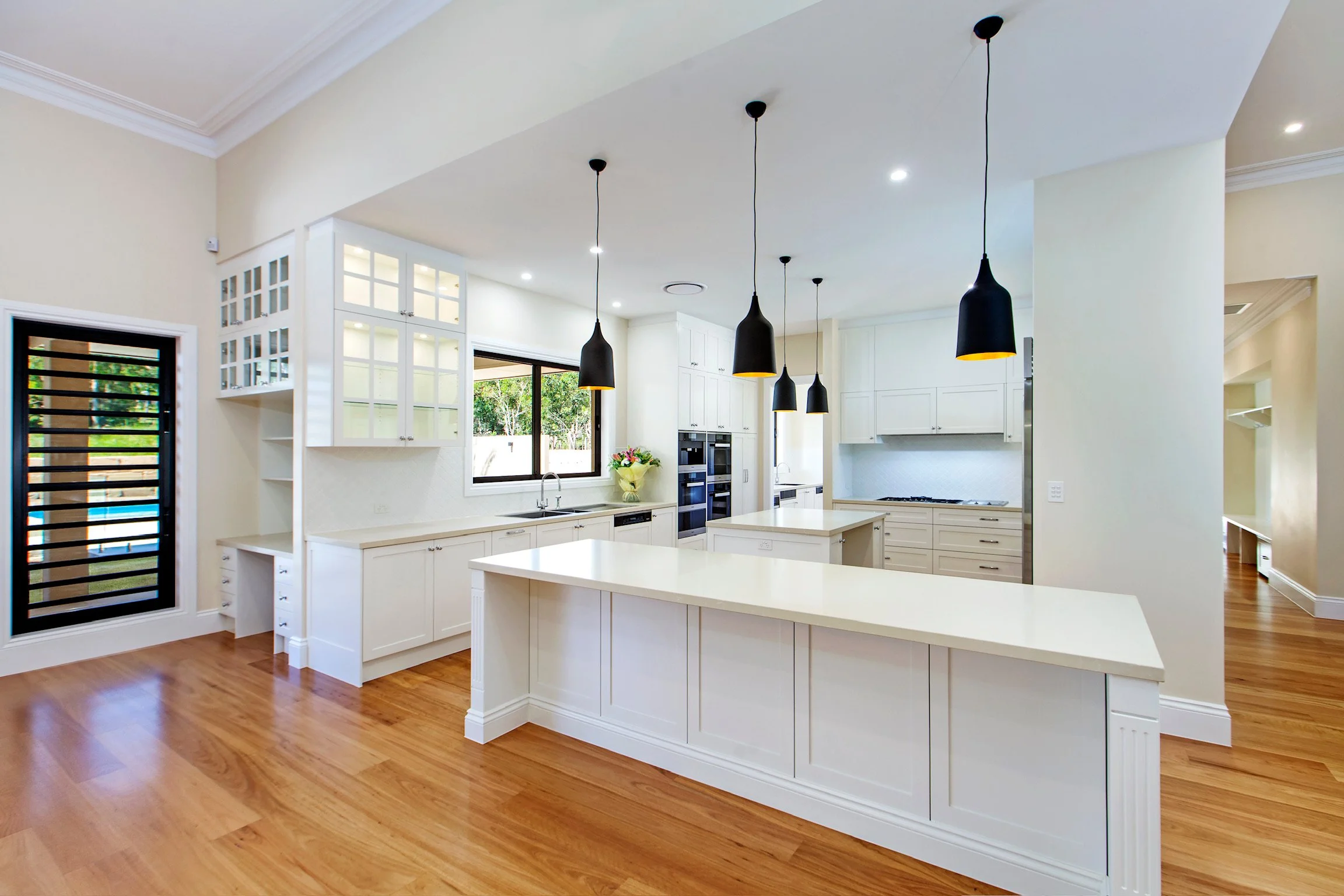 Bright modern kitchen with white cabinets, a large center island, black pendant lights, hardwood floors, and a window overlooking a backyard with greenery.