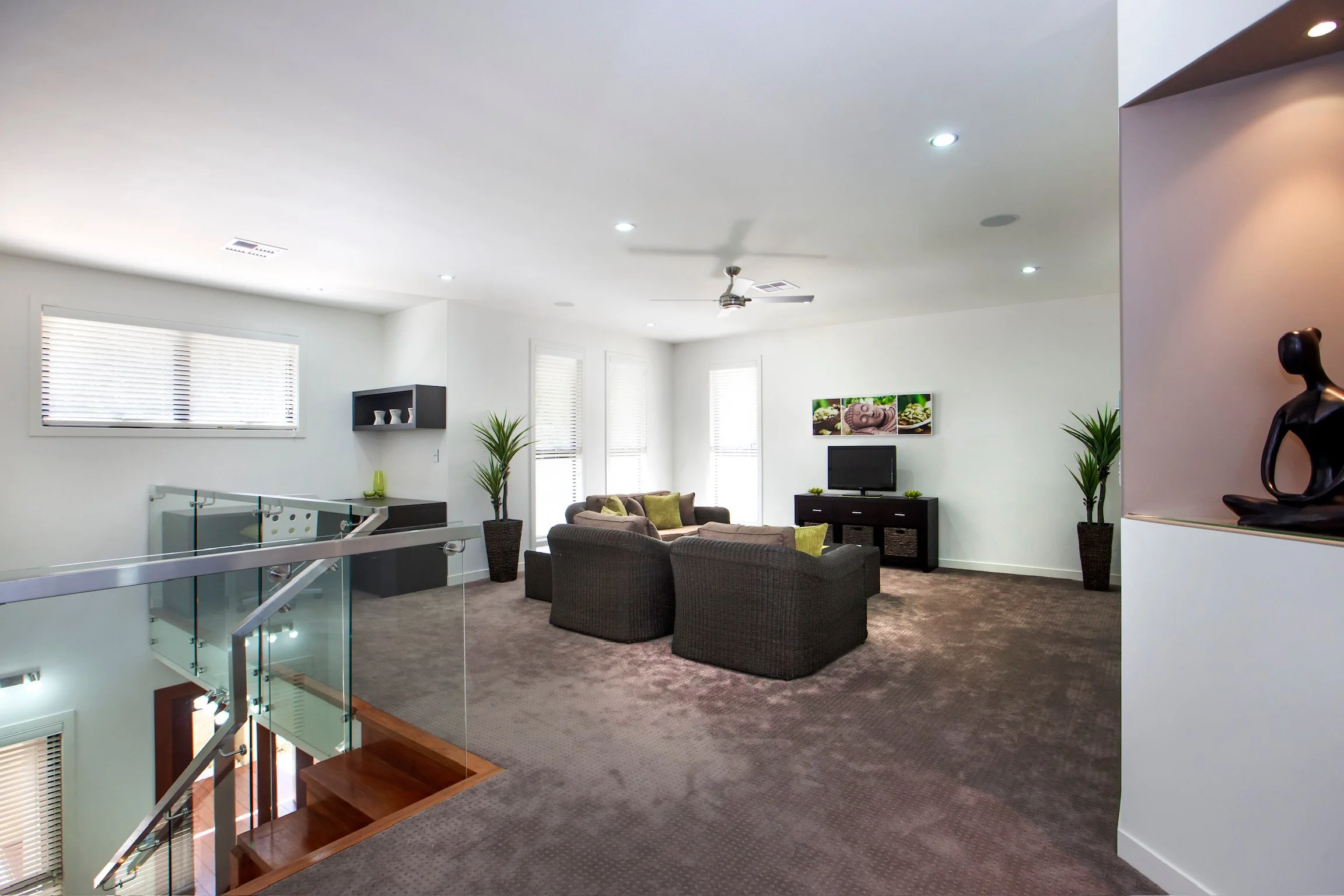 Living room with large windows, modern furniture including a sofa and armchairs, TV on a stand, potted plants, wall art, and a ceiling fan, with a staircase visible in the foreground.