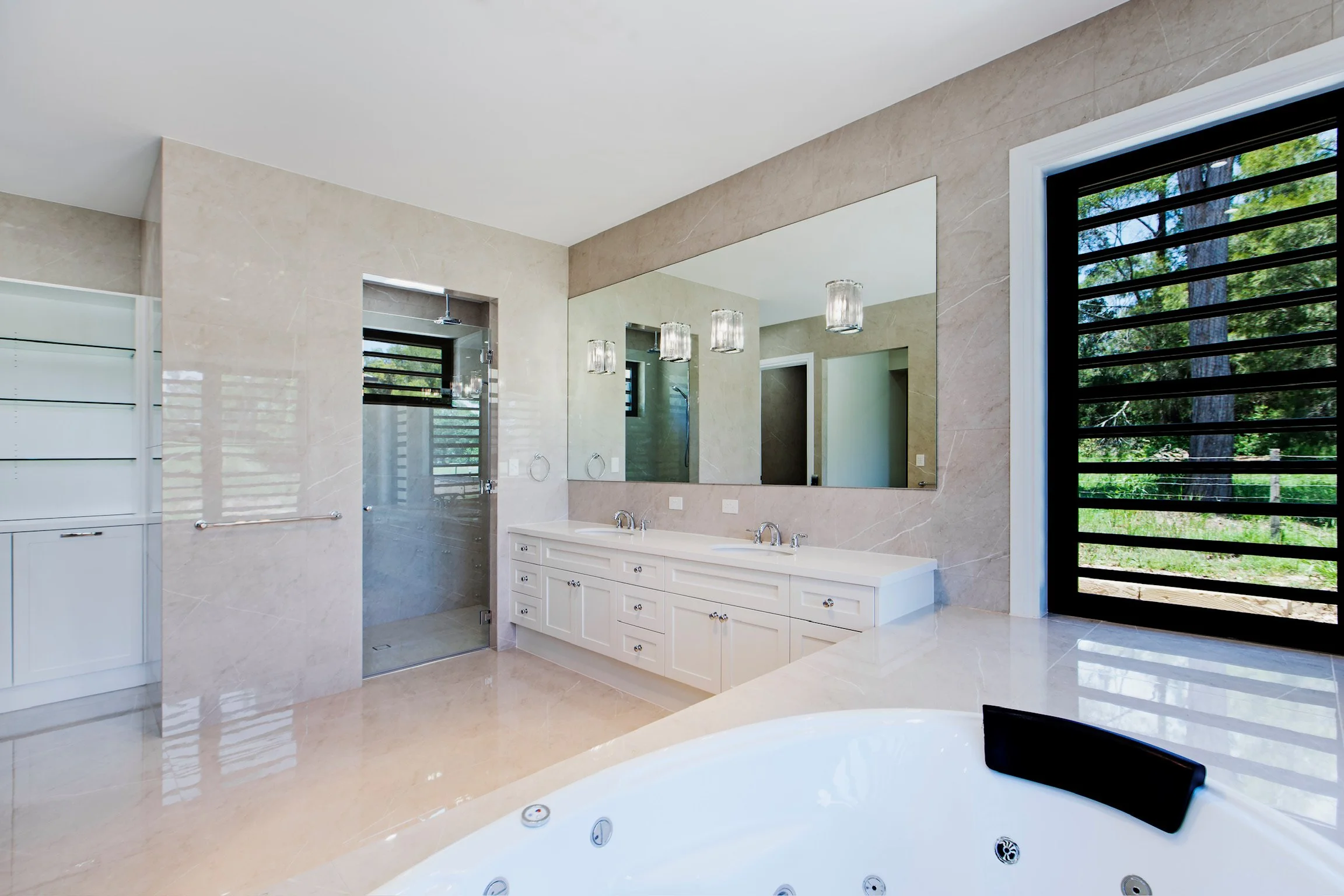 Modern bathroom with beige marble walls and floors, large mirror, double sink vanity, ceiling lights, a window with black metal bars showing outdoor trees, and a view of a spa bathtub in the foreground.