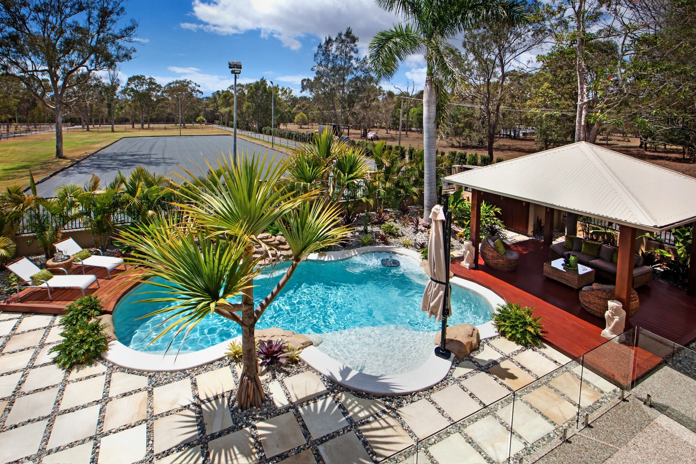 A backyard with a kidney-shaped swimming pool surrounded by a deck with lounge chairs, sun umbrellas, and lush tropical plants. In the background, there is a tennis court and trees under a partly cloudy sky.