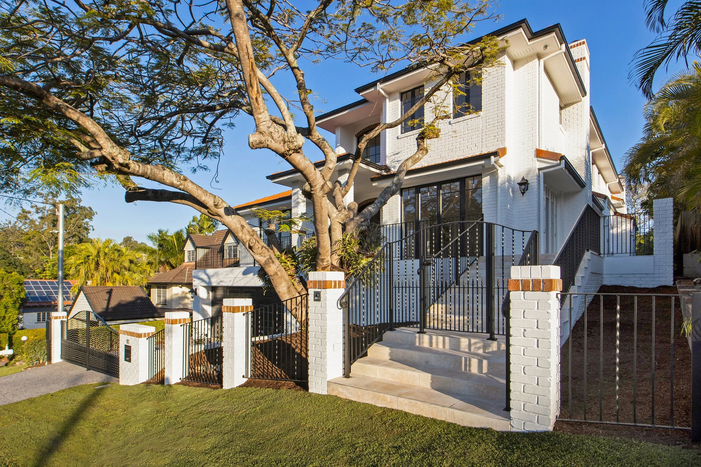 A two-story white house with black window frames, a black roof, and a front porch with stairs. A tall tree with spreading branches is in front of the house. The house is surrounded by a black metal fence with brick posts. The sky is clear and blue.