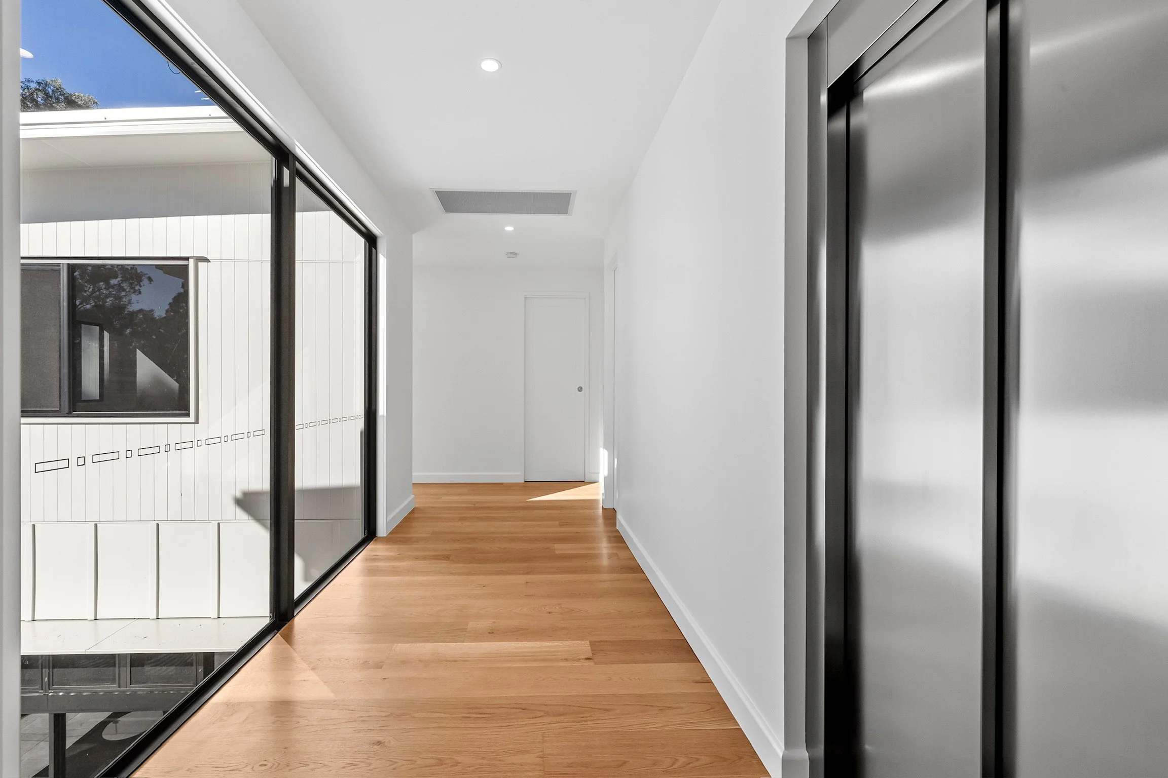 Modern hallway with white walls, hardwood flooring, and elevator doors on the right. Sunlight enters through large windows on the left, showing part of a neighboring building and trees outside.