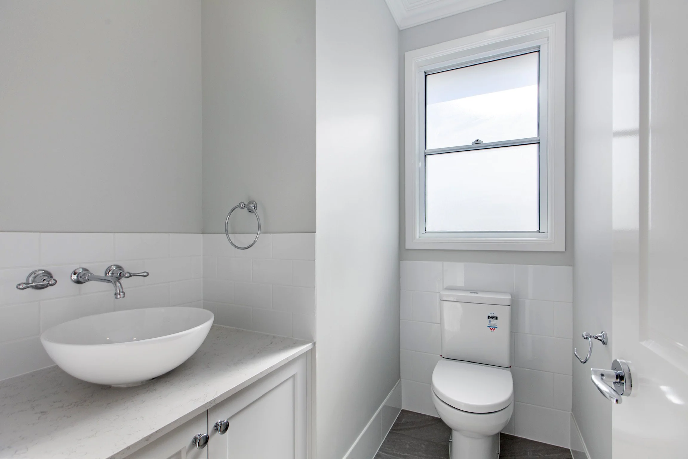 A clean, minimalist bathroom featuring a white ceramic vessel sink on a white countertop, chrome faucet, white tile walls, a small towel ring, a window with white trim bringing in natural light, a toilet, and a corner of a bathtub with chrome fixture