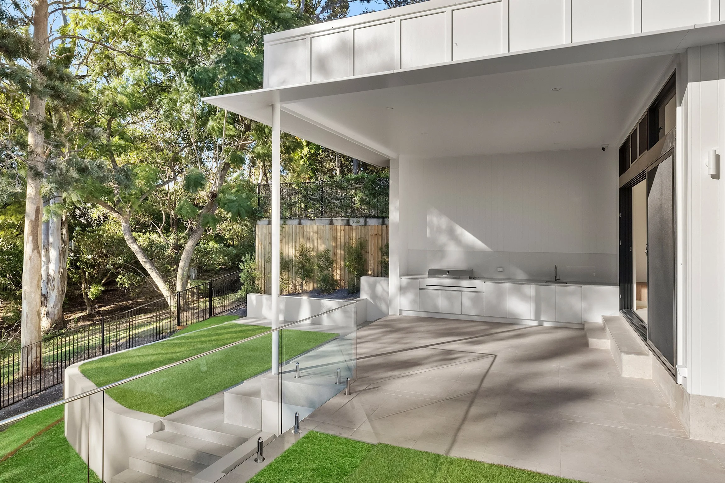 Modern house with an outdoor patio area featuring white cabinetry, glass railing, stairs, and a partial view of a backyard with trees and fence.