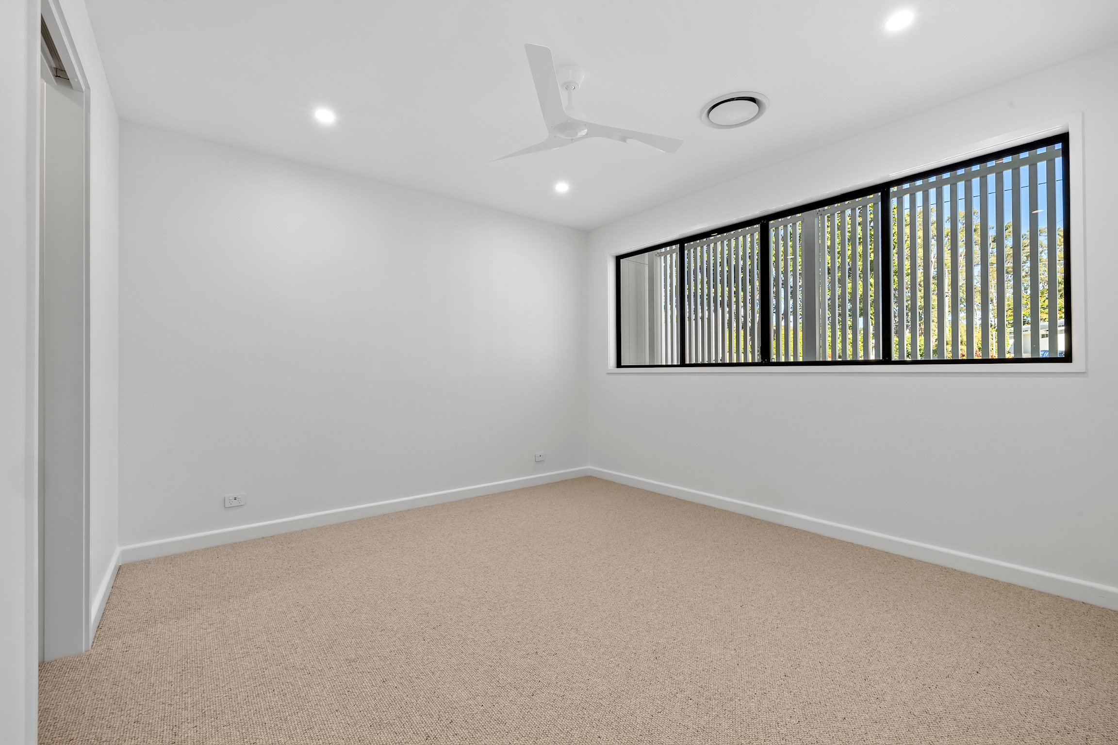 Empty room with beige carpet, white walls, large window with vertical blinds, ceiling fan, and ceiling lights.