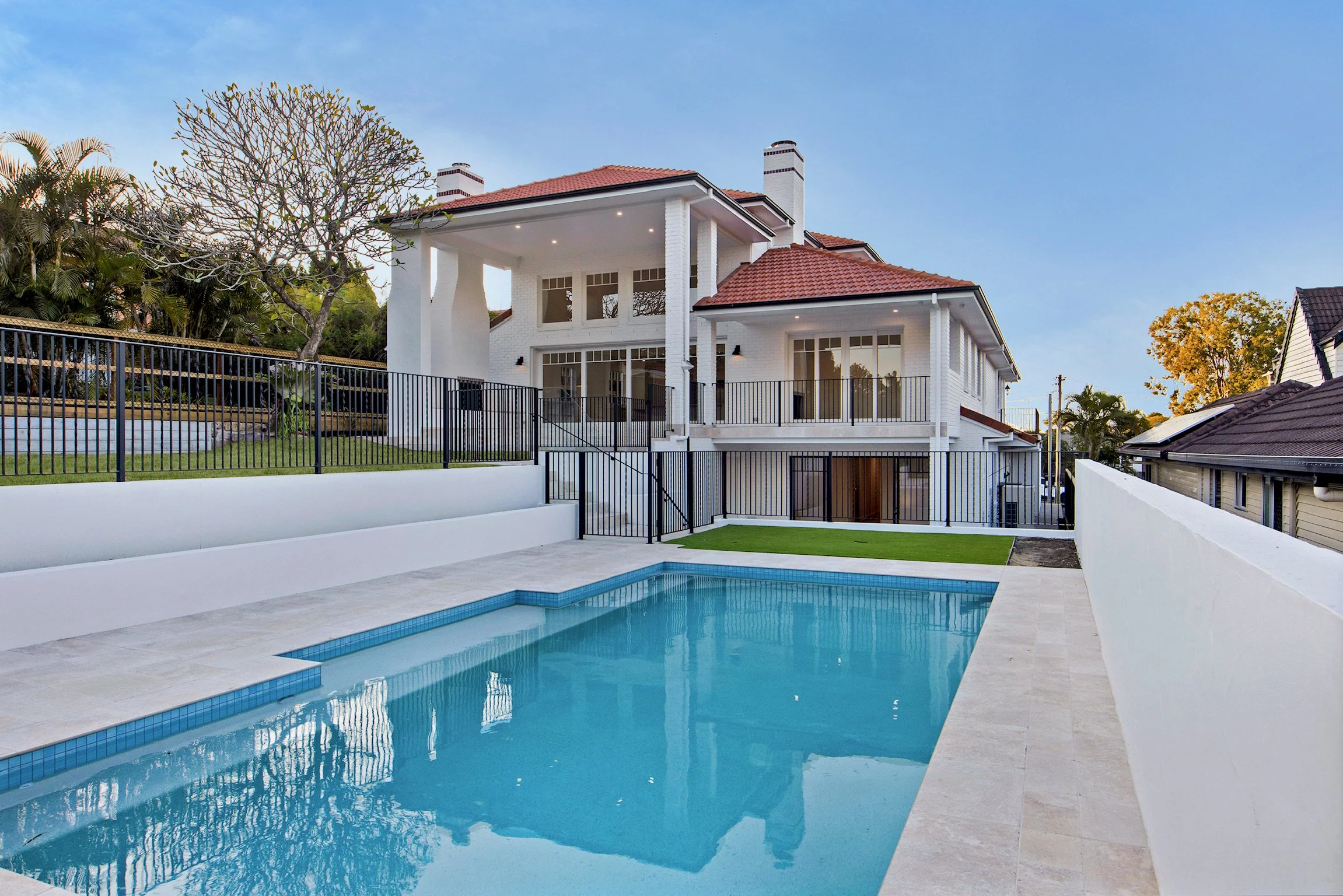 Modern white house with a red tile roof, multiple chimneys, and large windows, situated on a hill with trees, a black fence, and a backyard pool in the foreground during daytime.