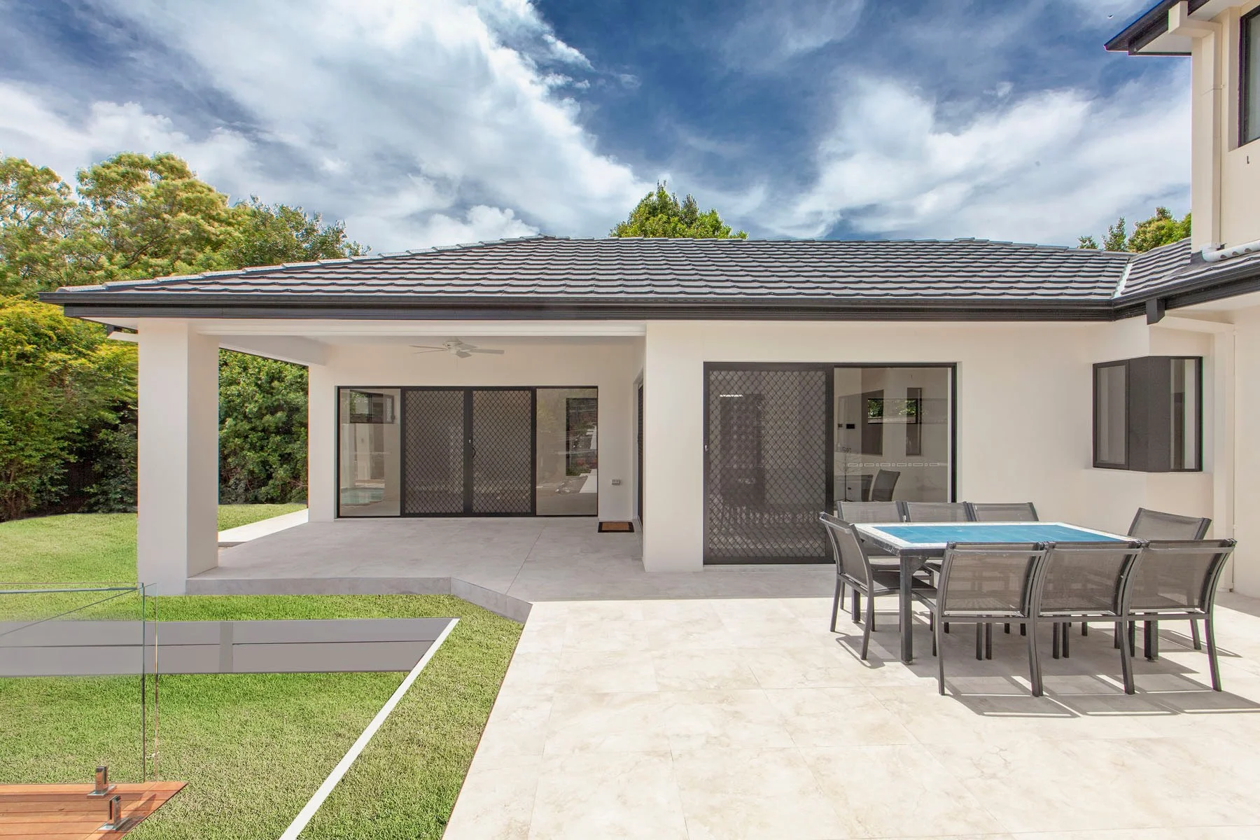 Modern backyard patio with dining table, chairs, sliding glass doors, and a lawn with a small fenced area, under a partly cloudy sky.