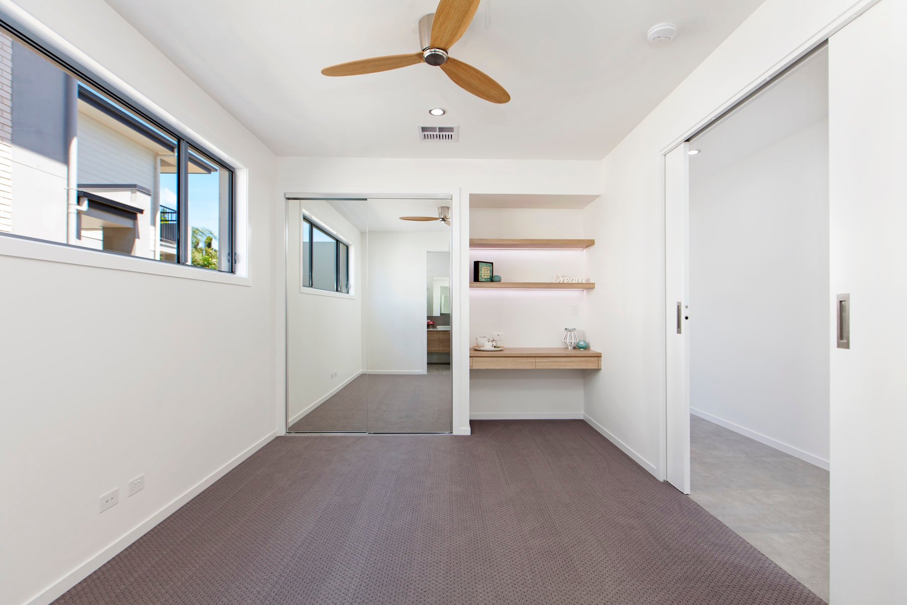 An empty room with white walls, a brown carpet, a ceiling fan, and sliding glass doors leading to another room with a similar style. There are open wooden shelves with decorative items and a small table in the background.