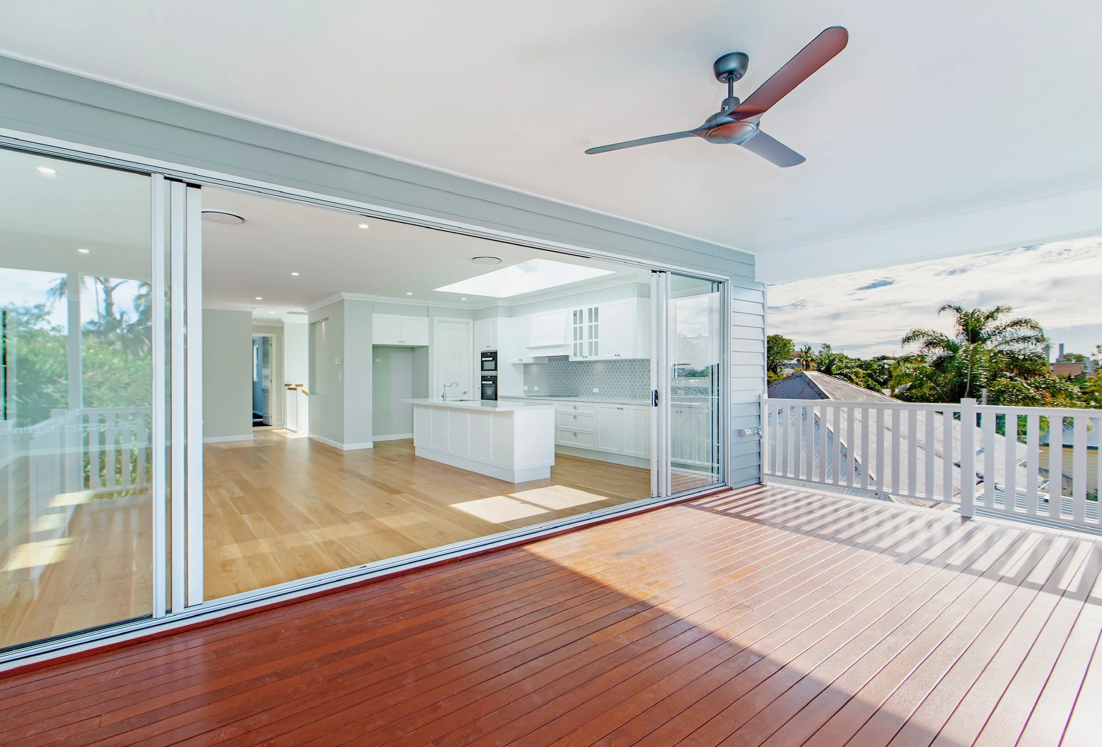 View of a spacious indoor-outdoor living area with a wooden deck, large glass sliding doors, and a balcony overlooking trees and houses, with bright natural light.
