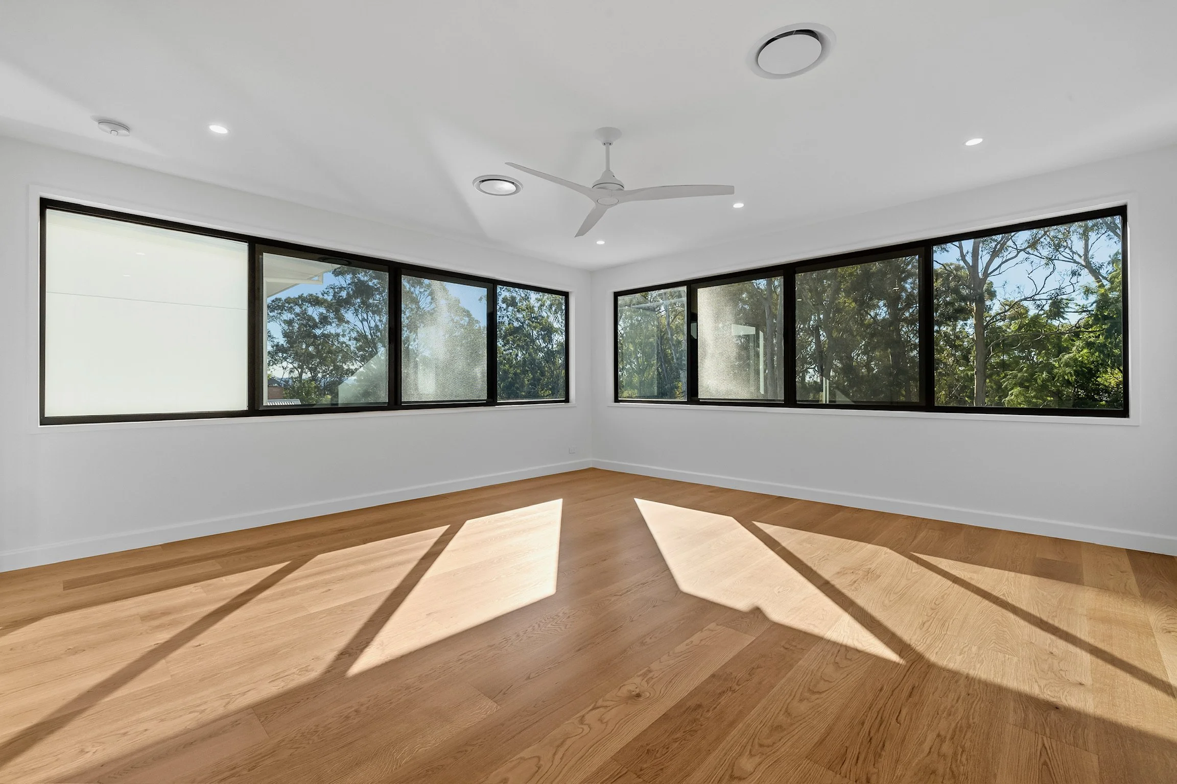 Empty room with large windows, white walls, hardwood floors, ceiling fan, and recessed lighting, with sunlight casting shadows on the floor.