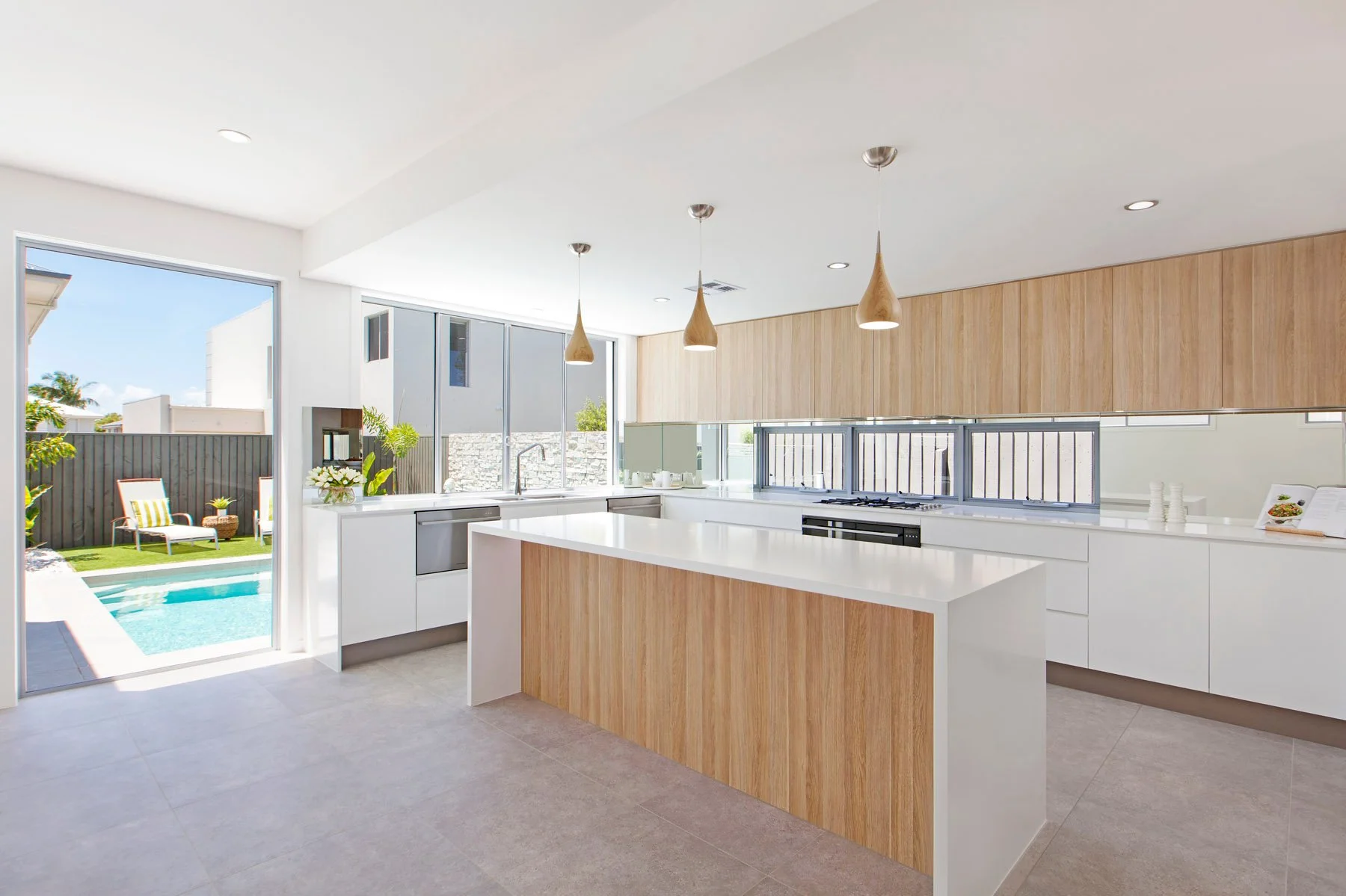 Modern kitchen with white cabinets, a wooden island, three pendant lights, large windows, and a view of a backyard with a pool, outdoor seating, and a fence.