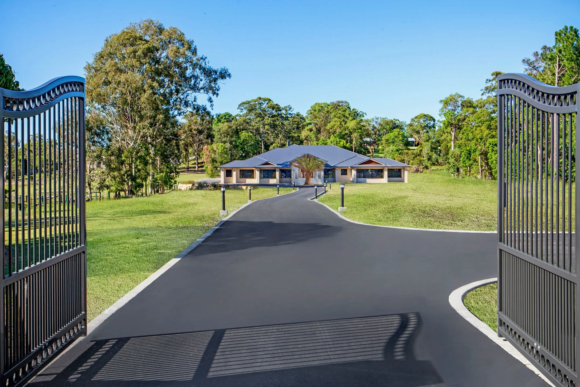 Open metal gates leading to a paved driveway that extends to a house in a green landscape with tall trees and a bright blue sky.