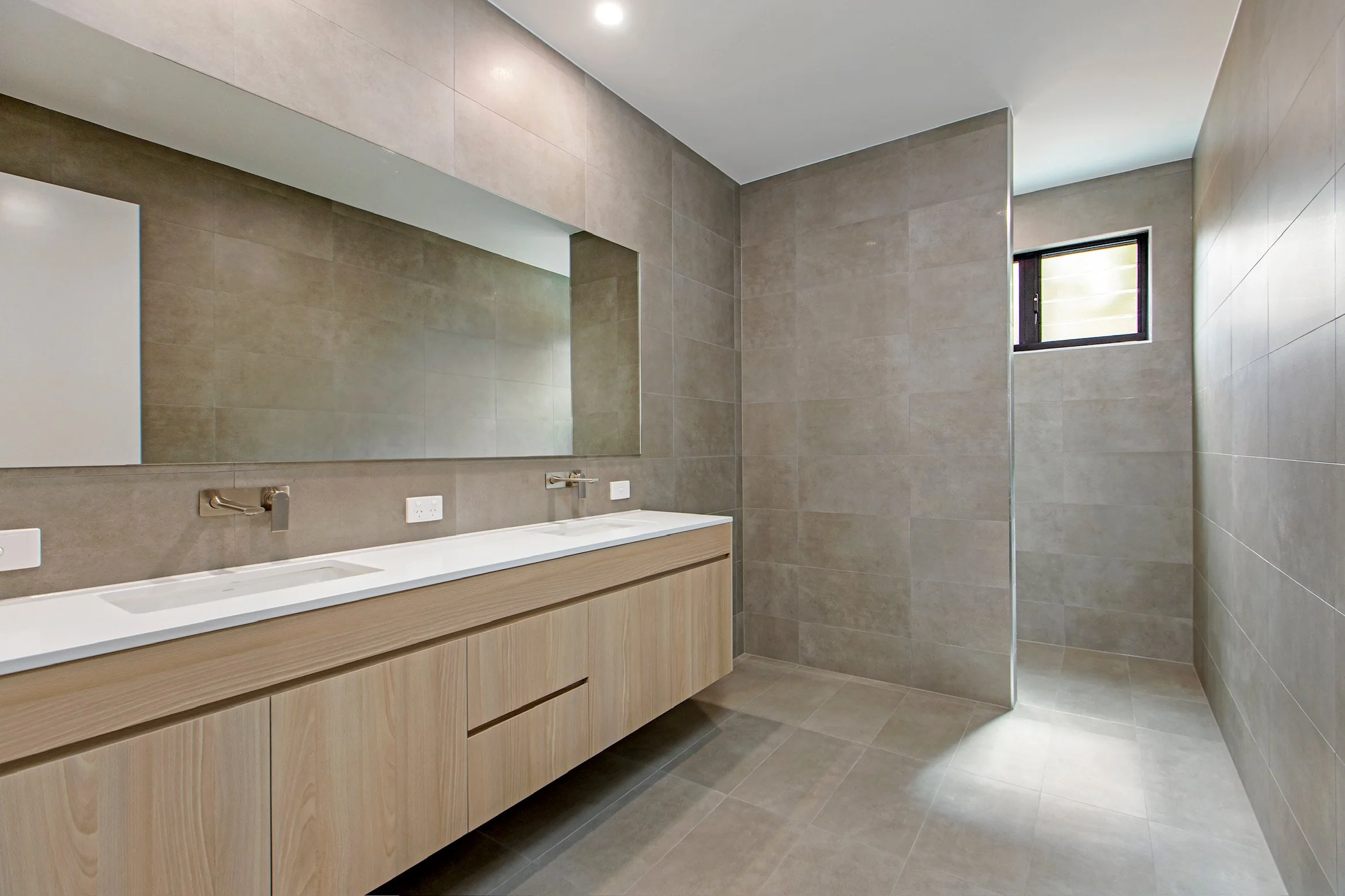 Modern bathroom with beige tiled walls and floor, a large mirror above a white counter with integrated sinks, a small window near the ceiling, and a walk-in shower area with glass partition.