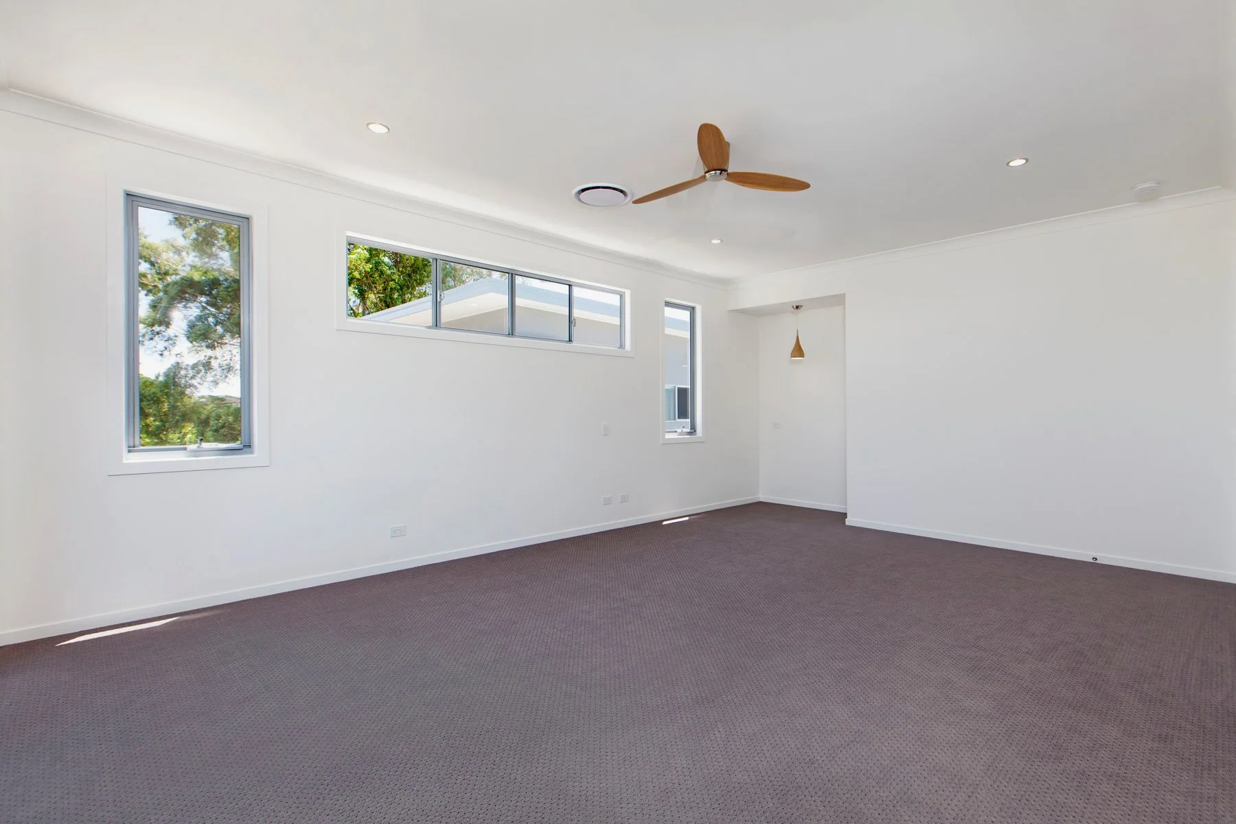 Empty living room with white walls, three windows, a ceiling fan, a pendant light, and brown carpeted floor.