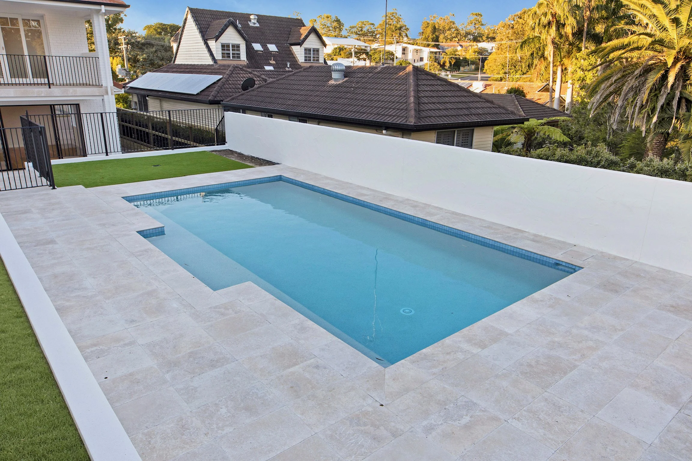 A backyard with a small swimming pool, a patch of artificial grass, and a white wall. In the background, there are rooftops of neighboring houses, palm trees, and trees under a clear blue sky.