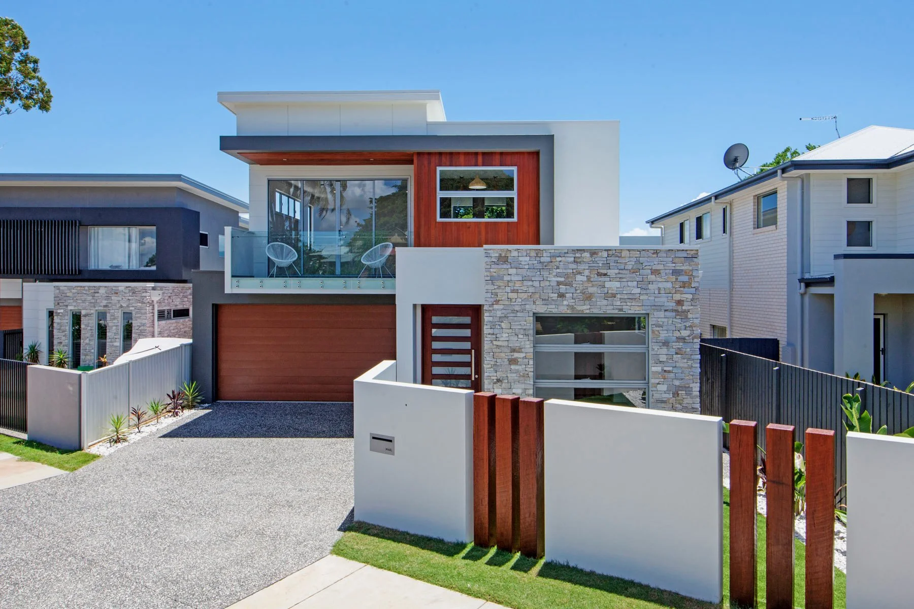Modern two-story house with a garden, stone and wood accents, a balcony with glass railing, and neighboring houses