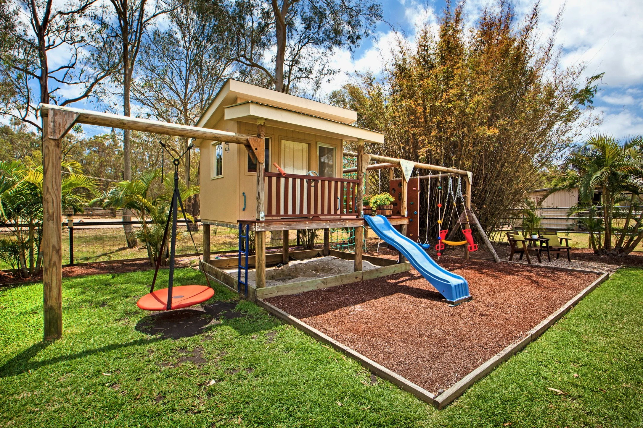 A backyard with a wooden playhouse, a slide, swings, and a sandbox surrounded by trees and plants.