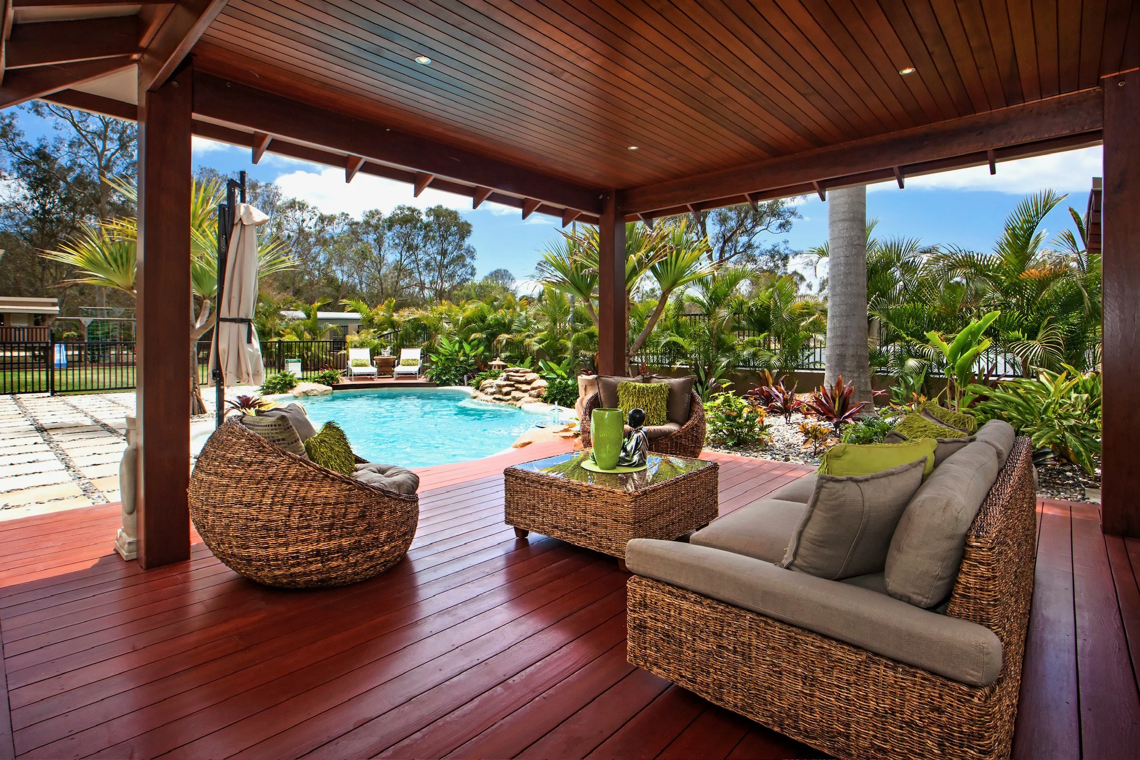 Covered patio with wicker furniture and cushions overlooking a backyard with a swimming pool, lush green plants, and trees.