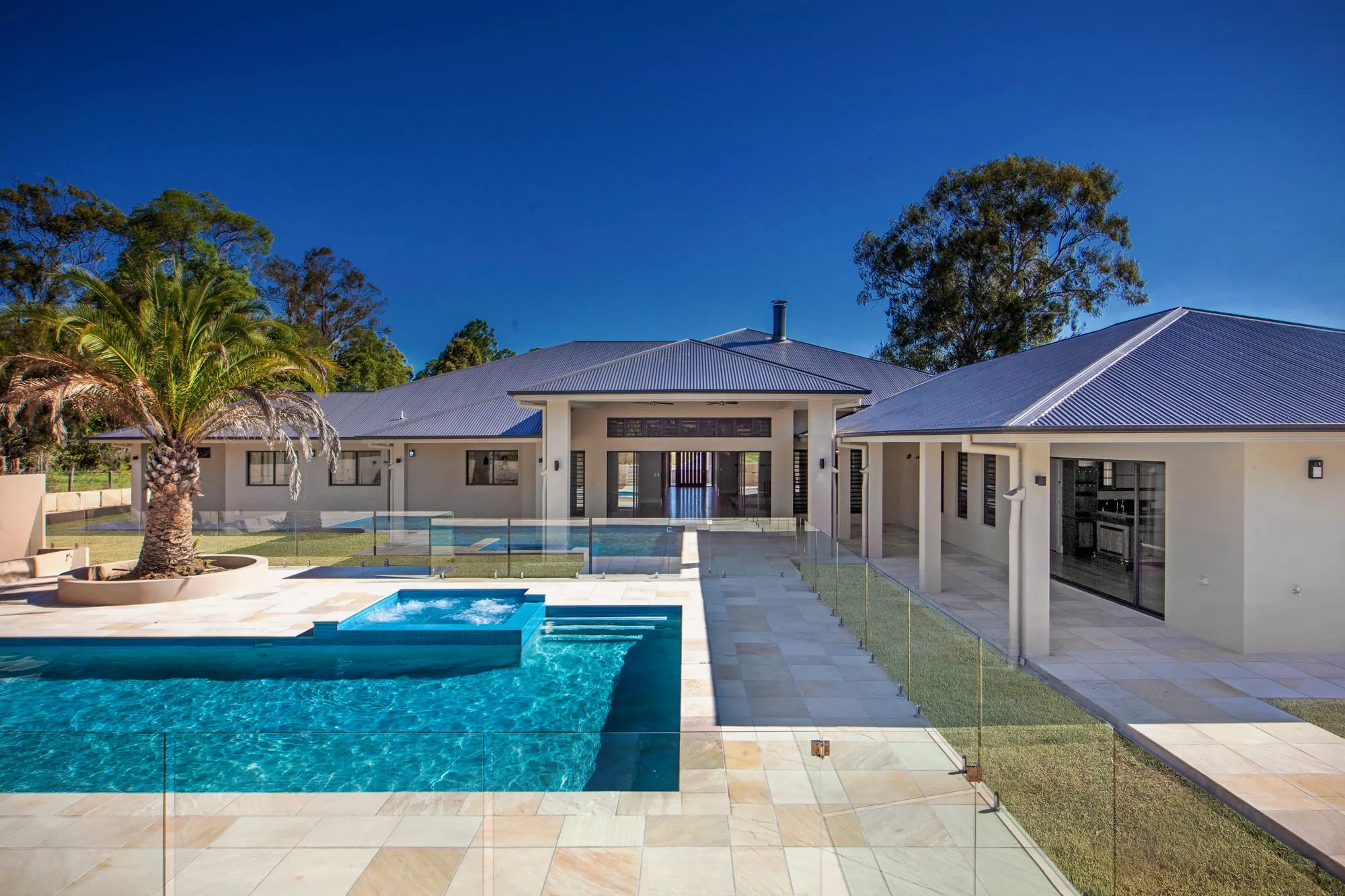 A modern house with a backyard that features a swimming pool, a small hot tub, a patio area, and tall trees in the background beneath a clear blue sky.