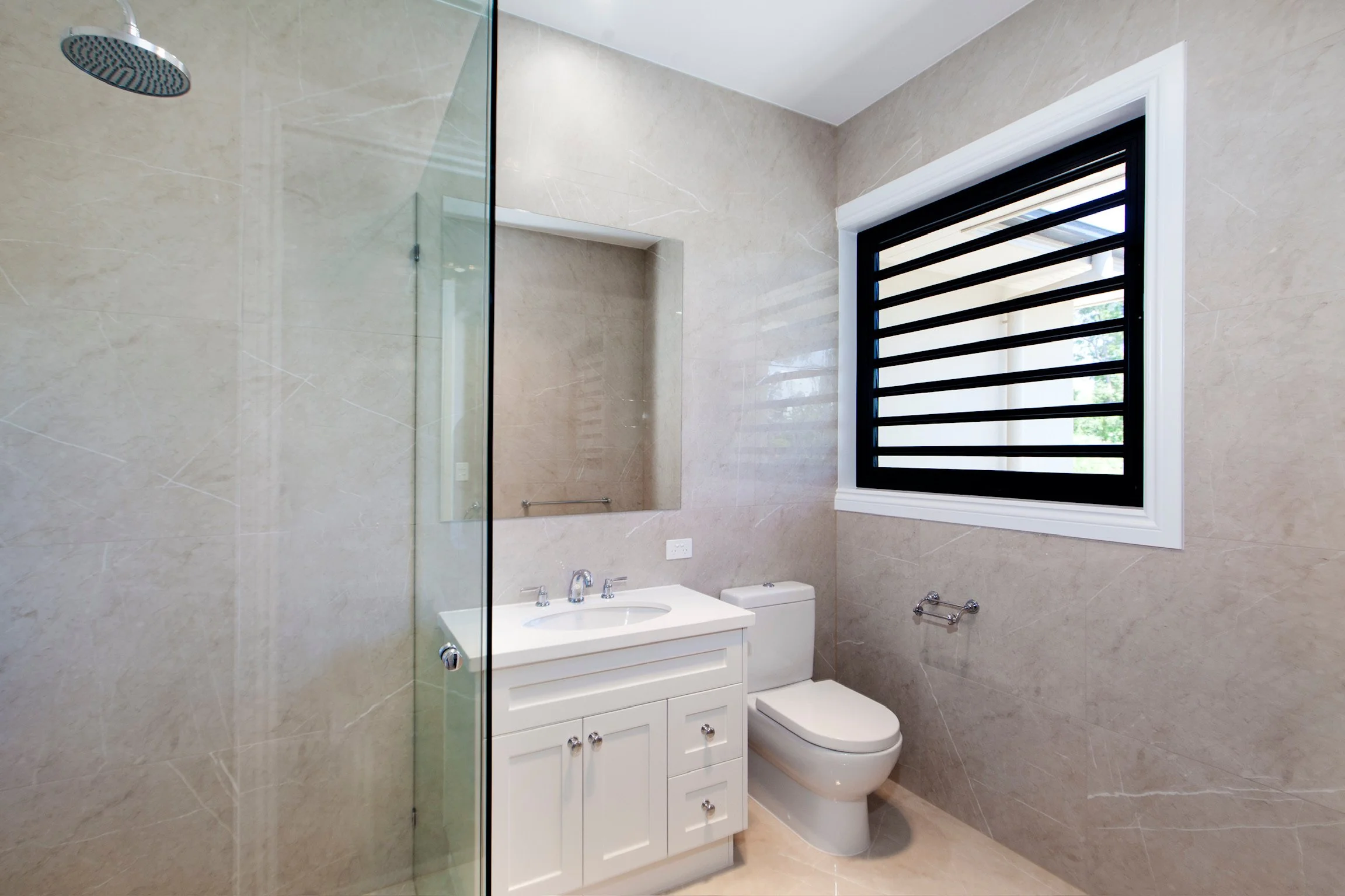 Modern bathroom with beige marble walls, a white vanity with sink and cabinet, a toilet, and a black-framed window with horizontal bars, natural light illuminating the space.