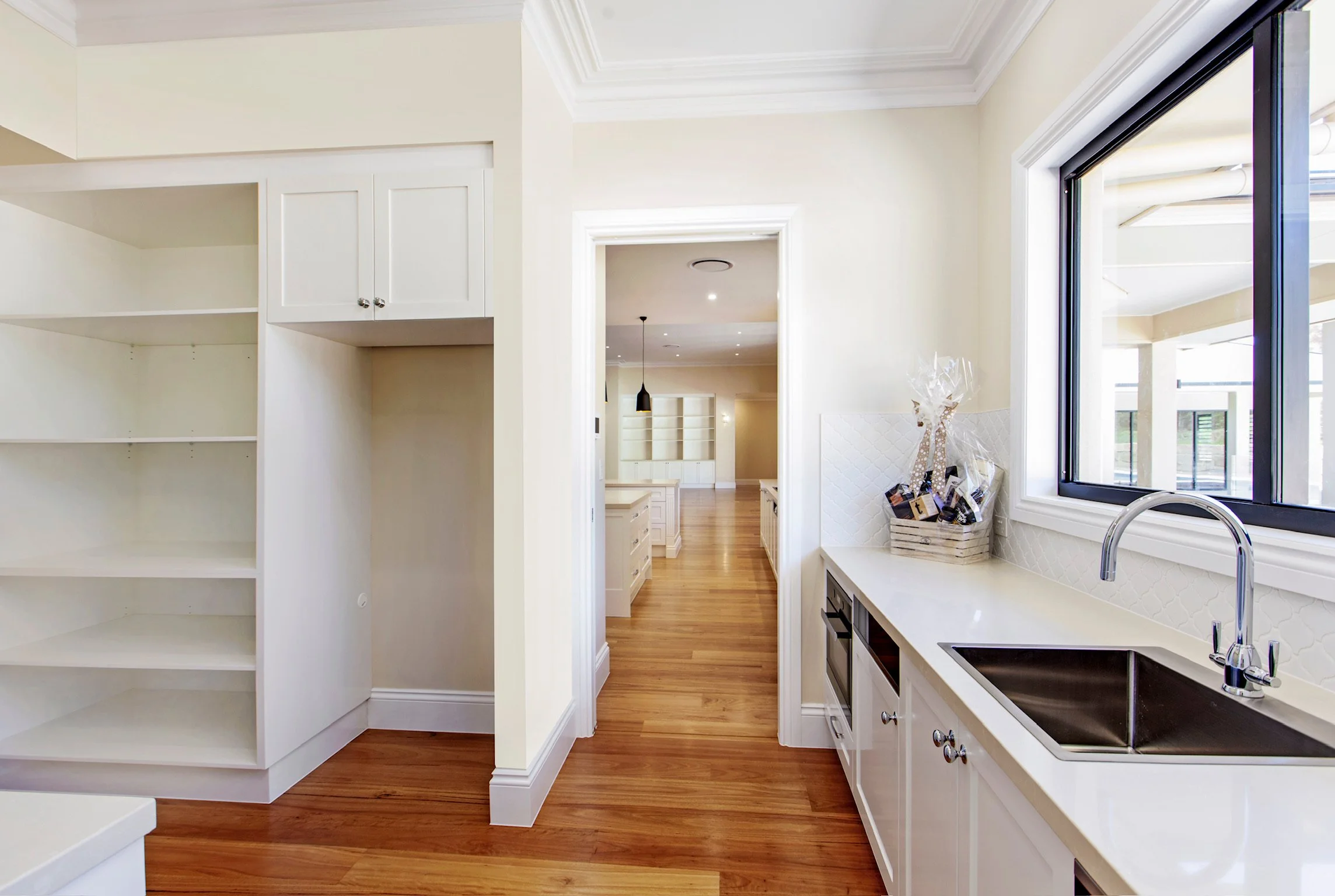 A bright, modern kitchen with white cabinetry, a large window above the sink, hardwood floors, and a view into another room with built-in shelves and pendant lighting.