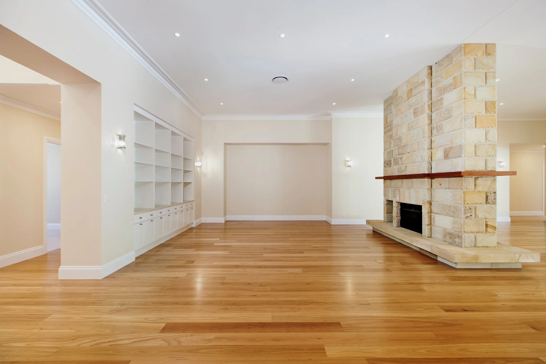 Empty living room with hardwood floors, white walls, a stone fireplace, white built-in shelves, and recessed ceiling lights.