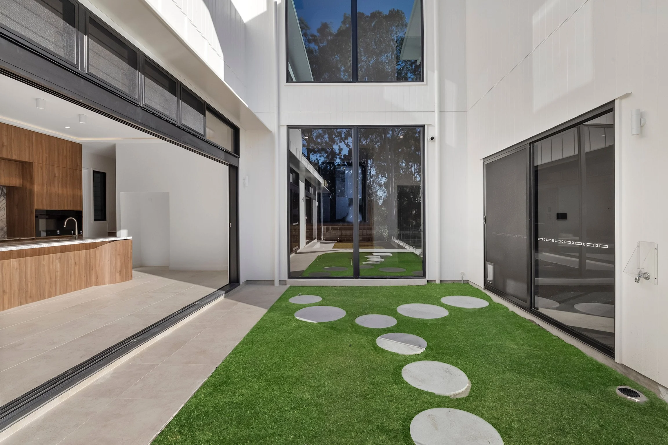 Modern home interior with a glass door opening to a small backyard with artificial grass and stepping stones, with large windows and a partial view of the kitchen.
