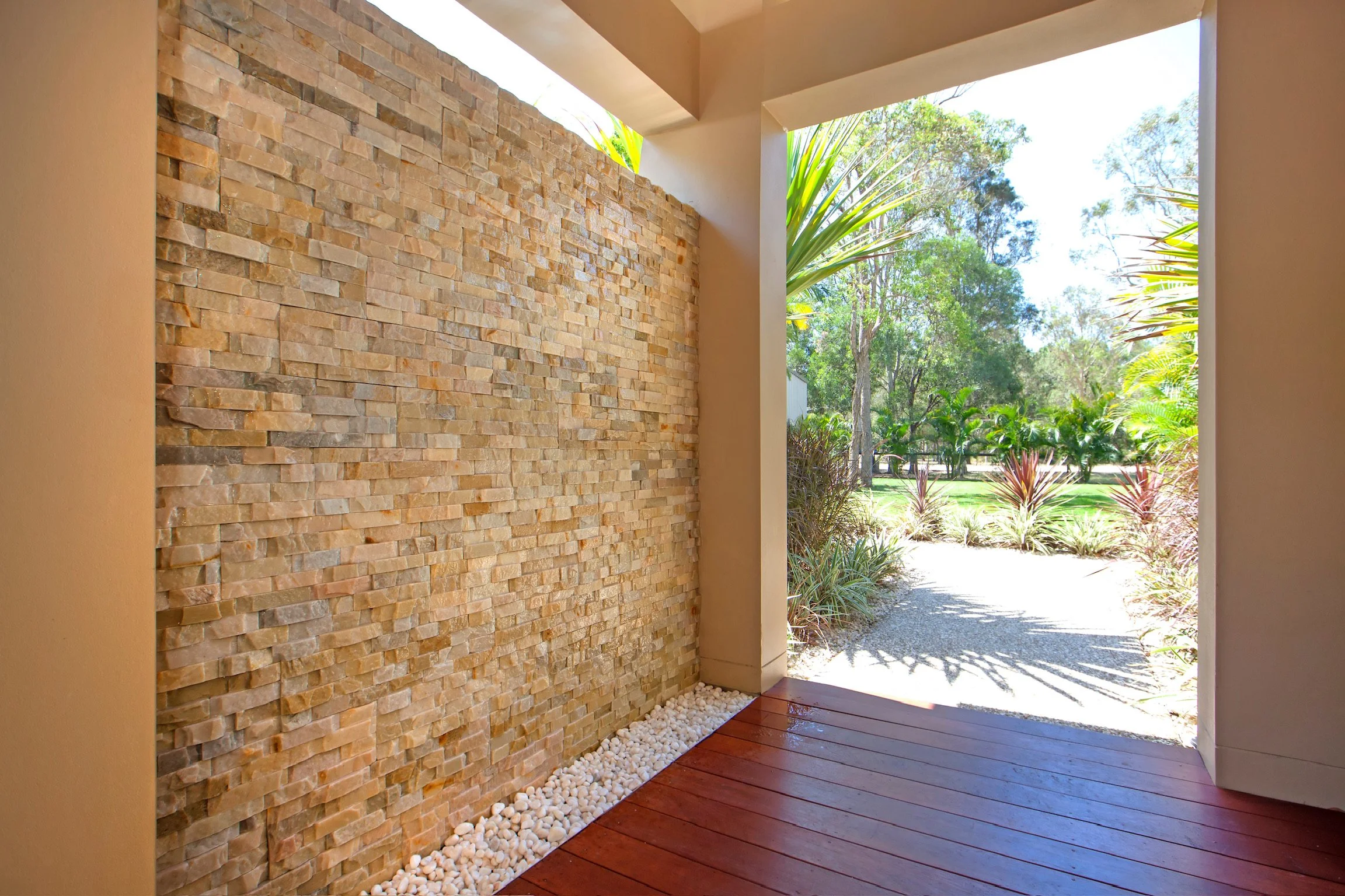 A porch with a textured stone wall on the left, a wooden floor, and an open view to a garden with trees and plants.