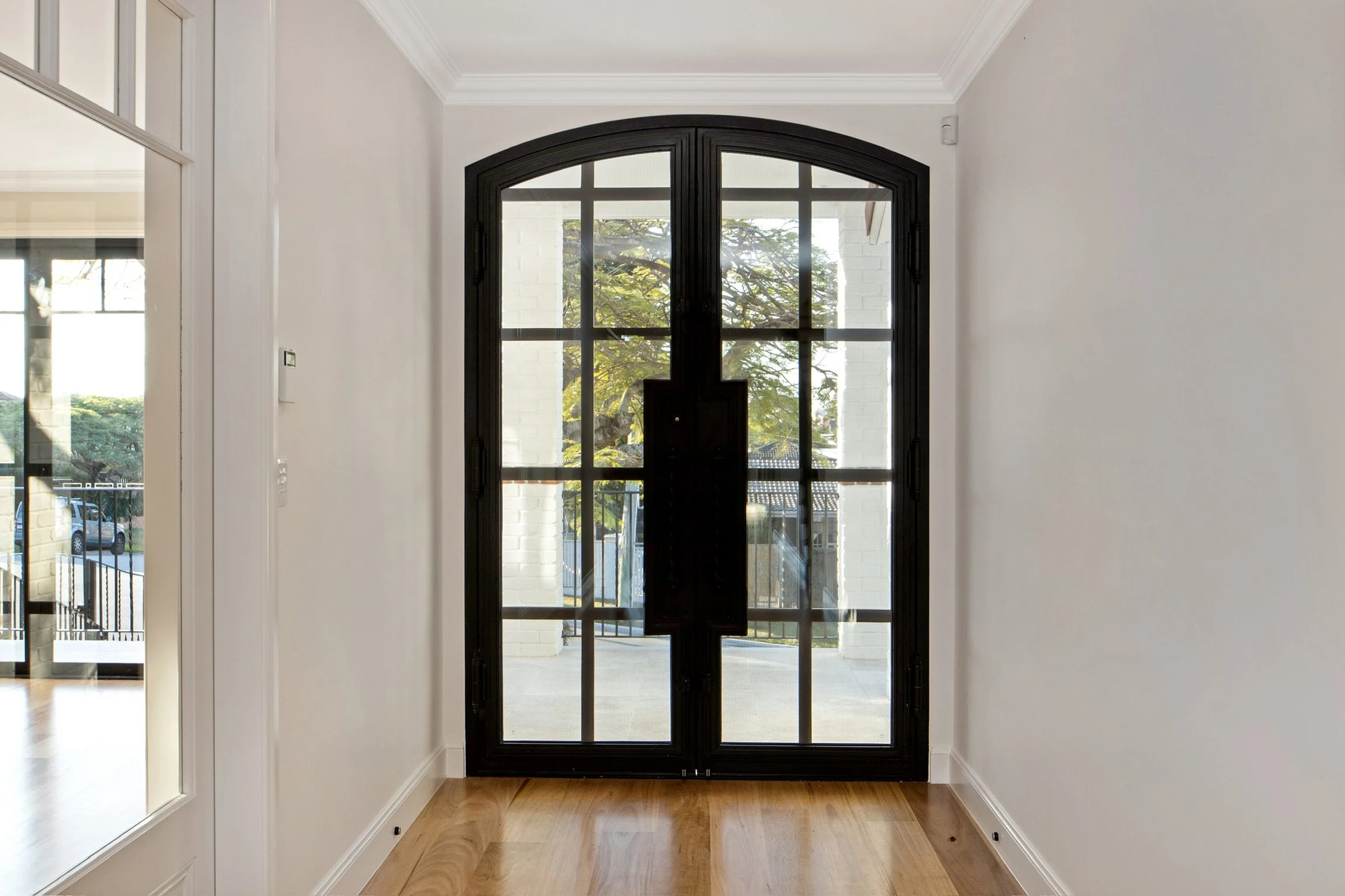 Interior view of a modern entryway with a large black framed glass door, white walls, crown molding, and wooden flooring.