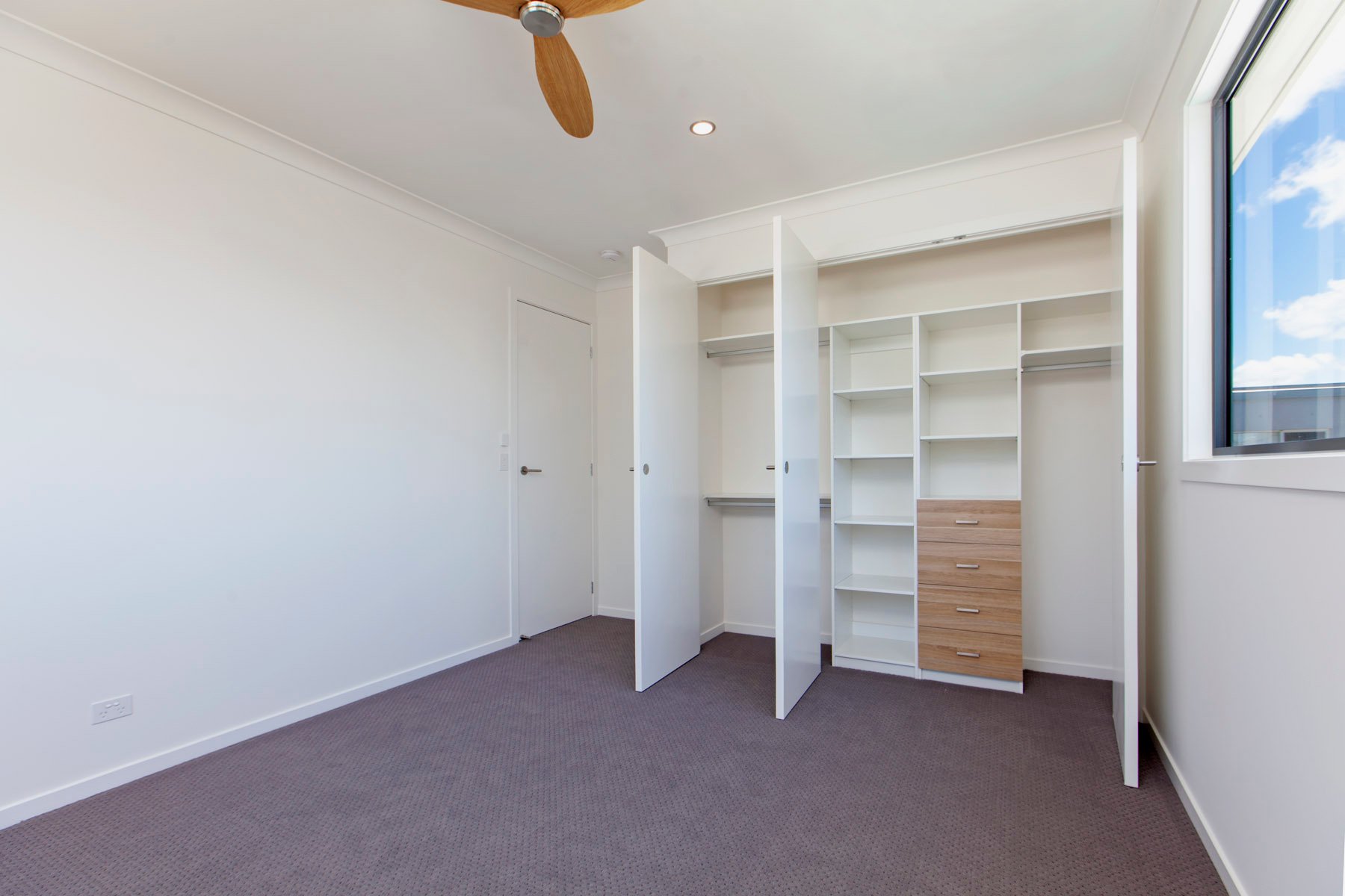 Empty walk-in closet with white built-in shelves and drawers, a window with blue sky and clouds, and a ceiling fan.