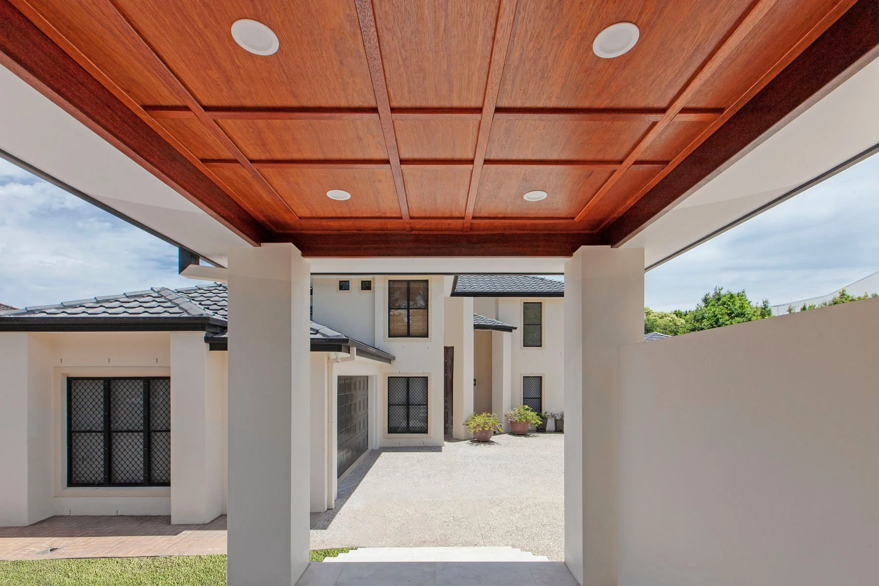View of a modern house exterior with a covered porch featuring a wooden ceiling with built-in lights. The house has multiple windows with black security screens, a garage door, potted plants, and a concrete driveway. The house is surrounded by white 