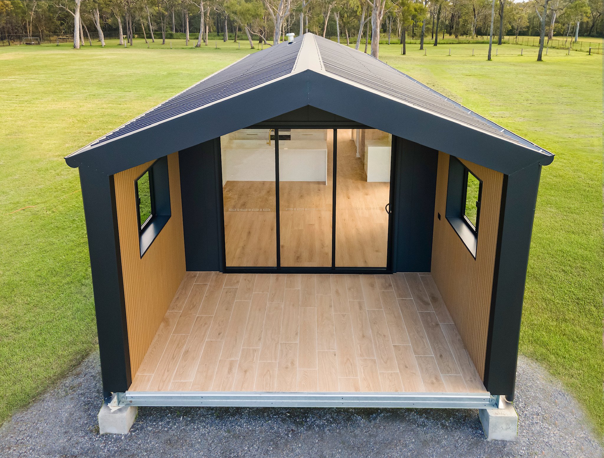 A partially assembled tiny house with large glass sliding doors and a wooden floor, situated on a grassy area with trees in the background.