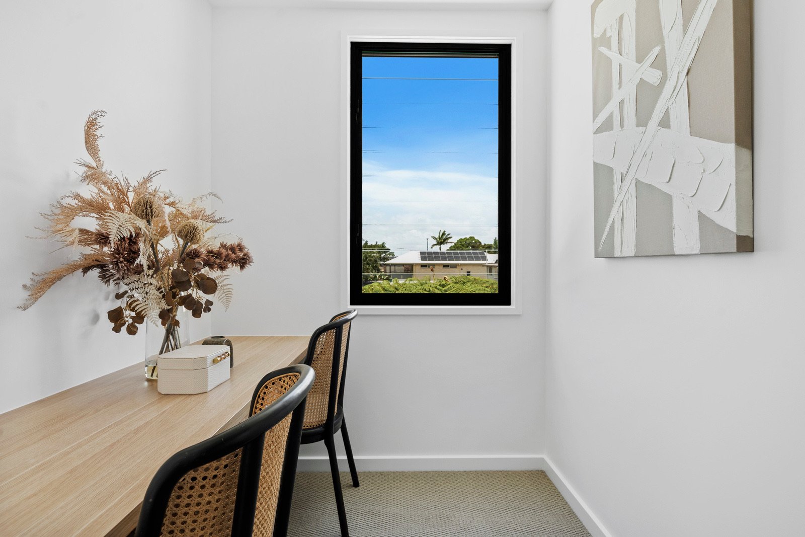 Interior of a minimalistic dining room with a rectangular wooden table, black chairs with woven seats, a vase of dried flowers, a white box, a window showing a sunny day, white walls, and abstract wall art.