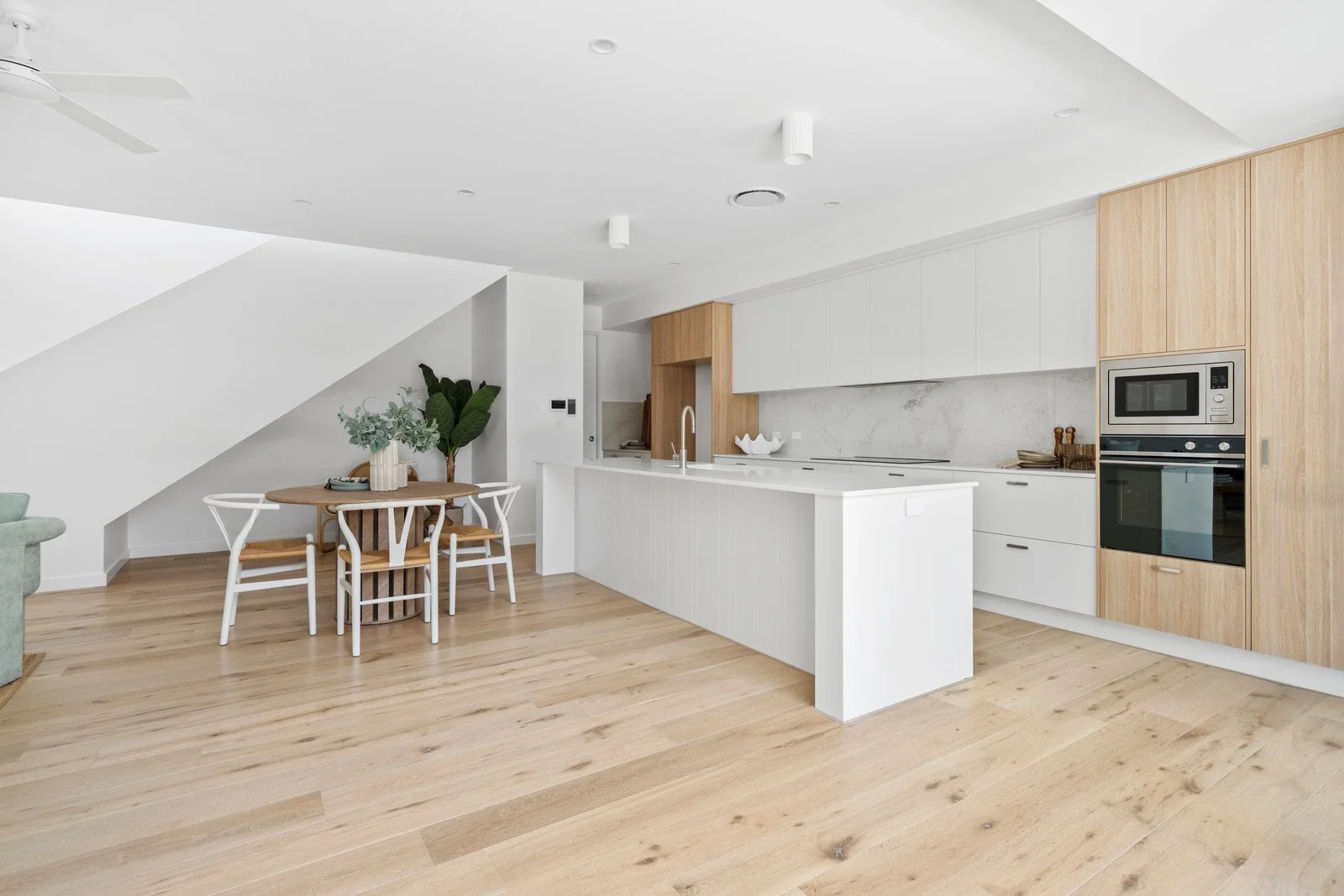 Modern kitchen with white cabinets, a kitchen island with a sink, built-in microwave and oven, and a small dining area with a round wooden table and four white chairs with wooden seats. Light wood flooring, a few decorative plants, and minimal decor.
