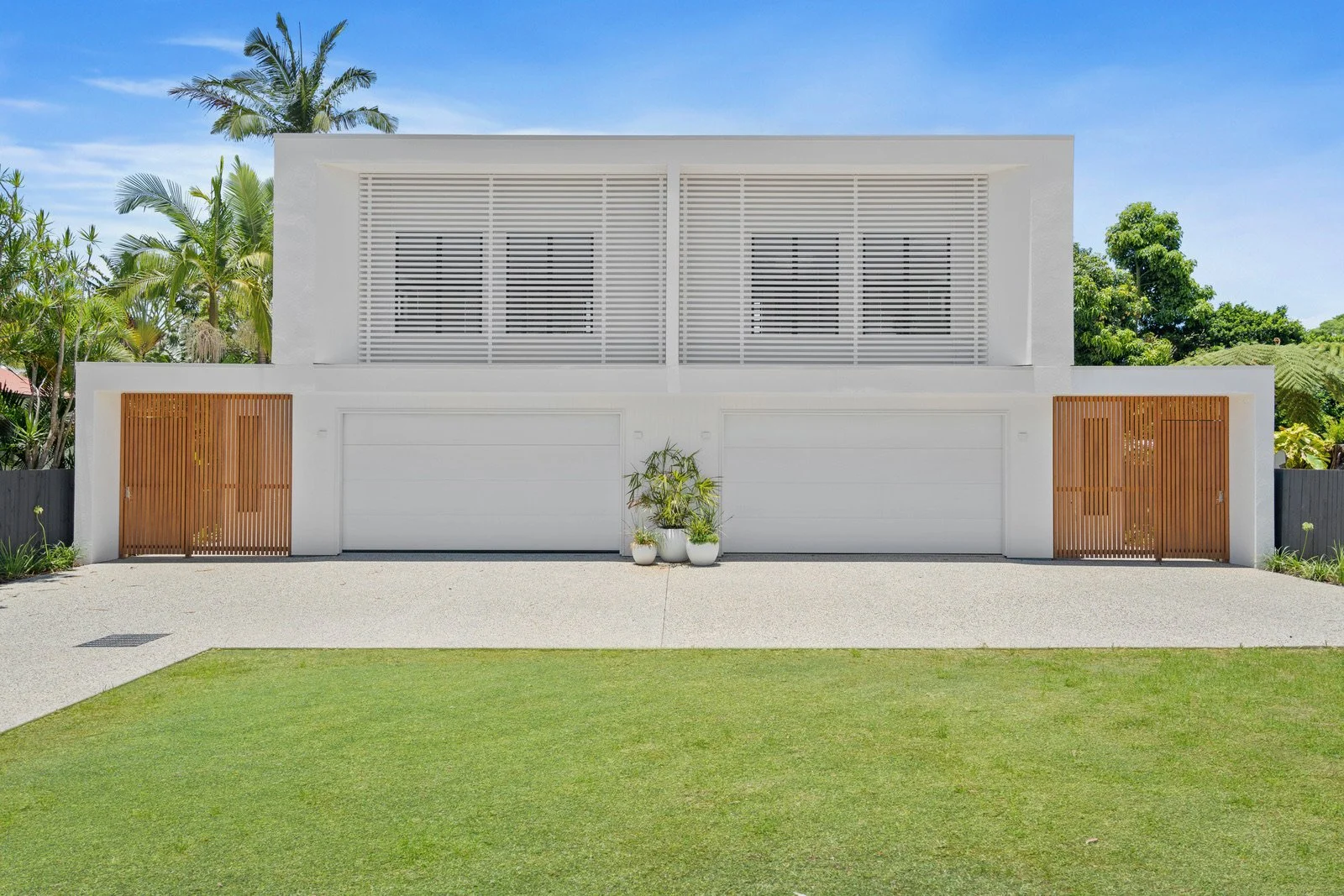 Modern white house with two garage doors, wooden accents, and potted plants in front, surrounded by greenery and clear sky.