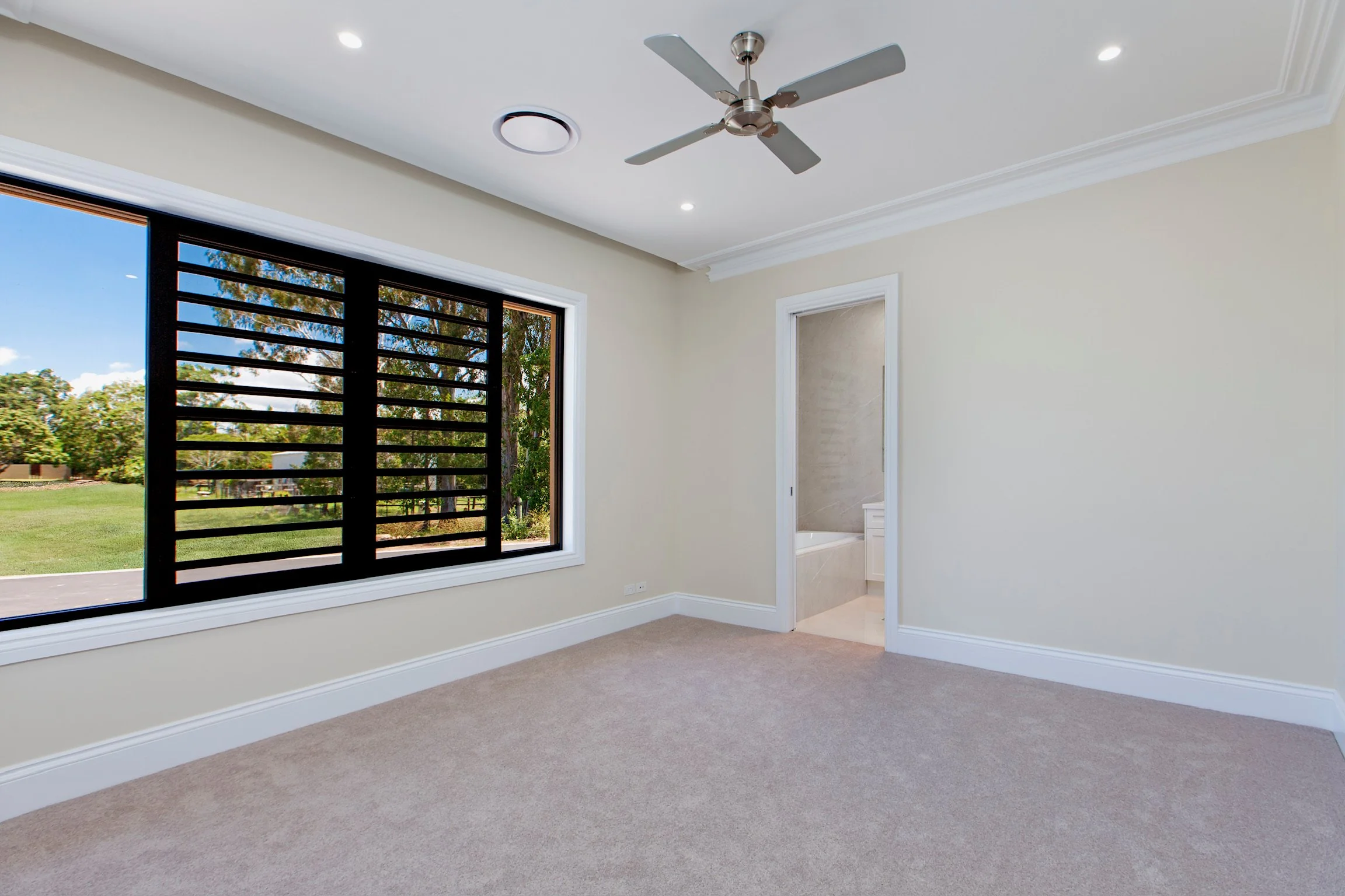 Empty room with large window, white walls, beige carpet, and ceiling fan.
