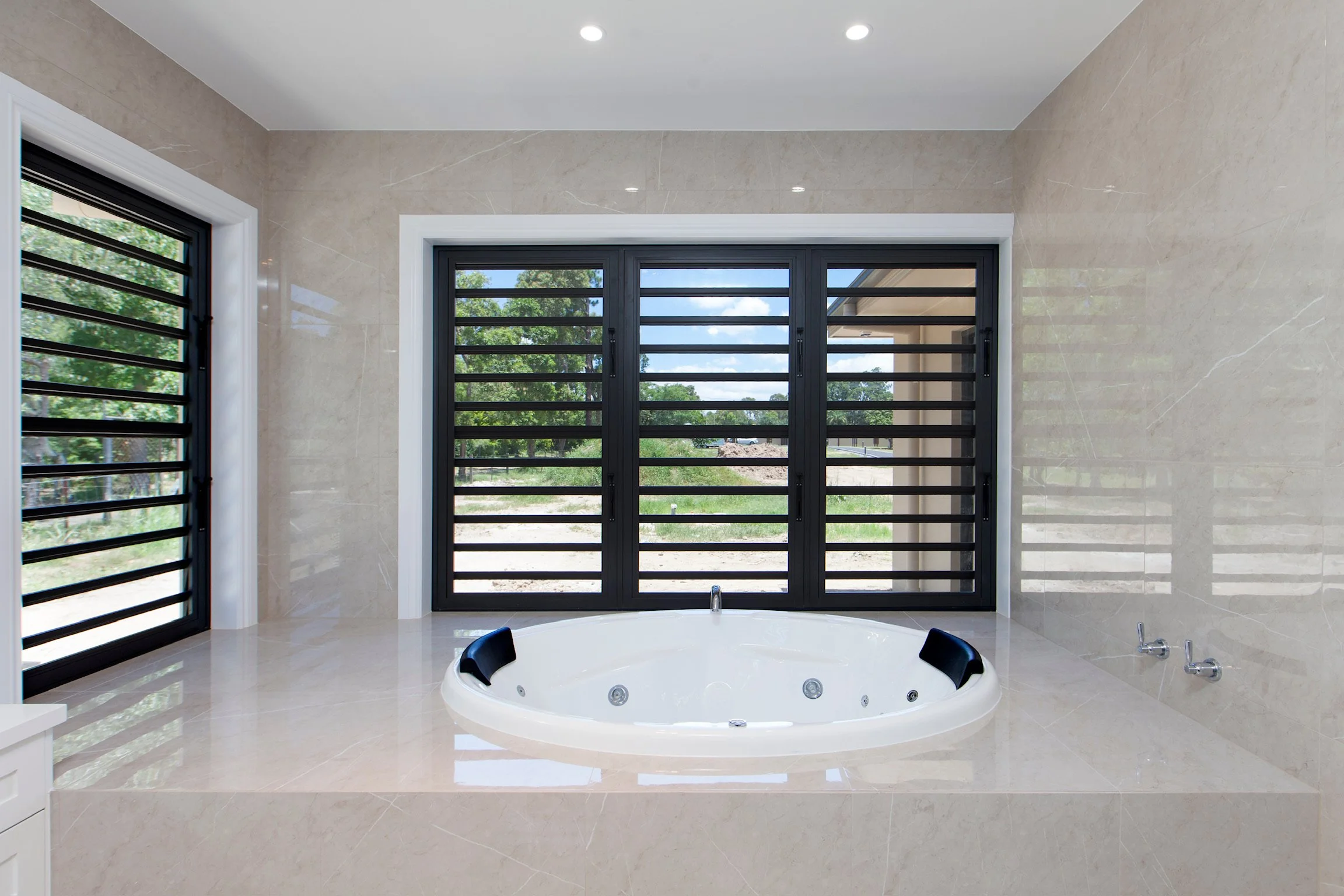 Bathroom with beige tiled walls and floor, large window with black bars showing trees outside, and a white whirlpool bathtub with black headrests.