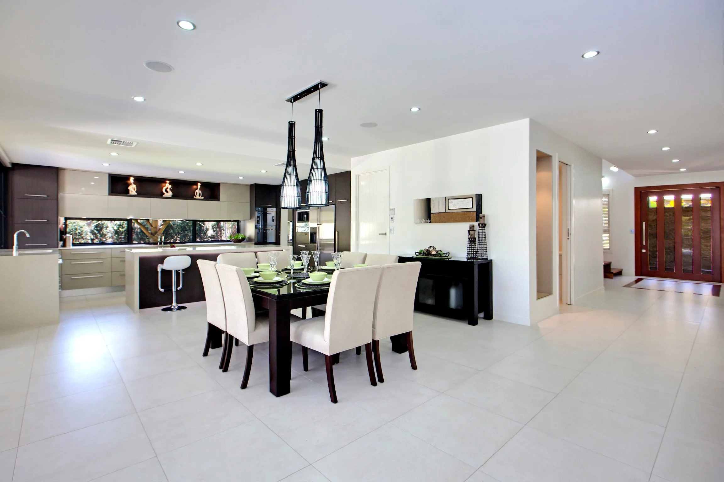 Modern kitchen and dining area with a black dining table set with bowls and glasses, cream-colored chairs, black pendant lights, white cabinetry, and large windows.