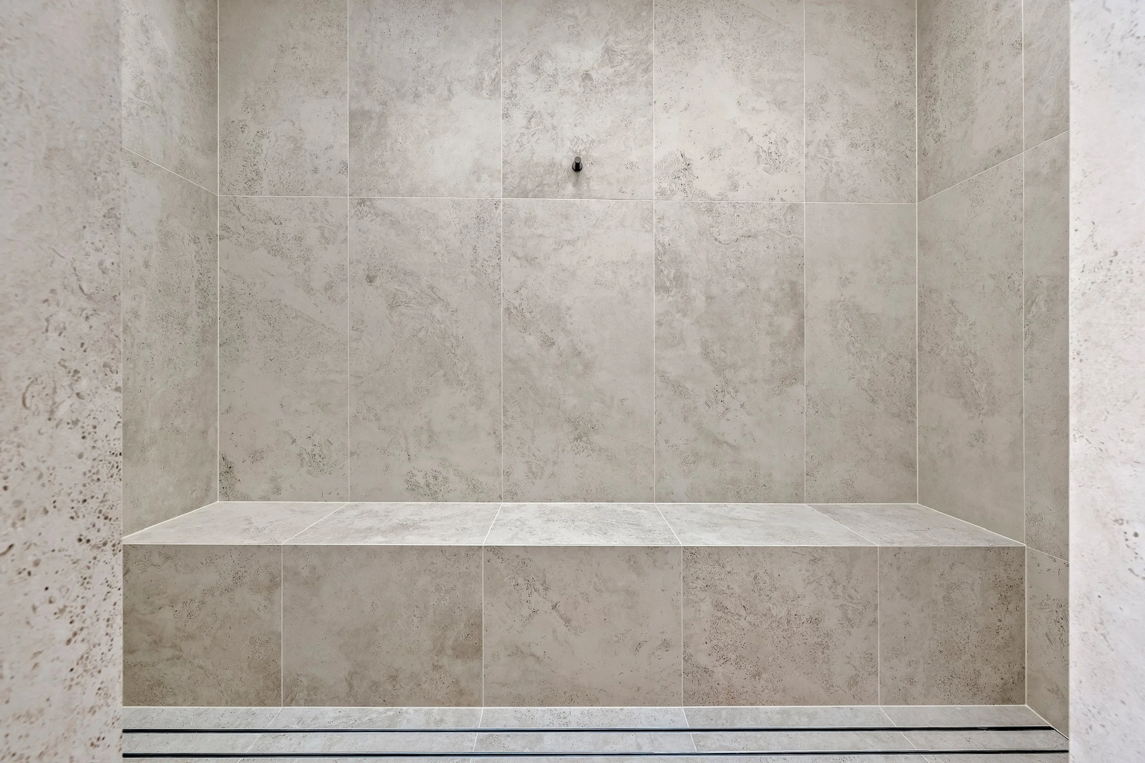 Empty shower with light gray textured tile walls and floor, with a small shower head in the center of the back wall.