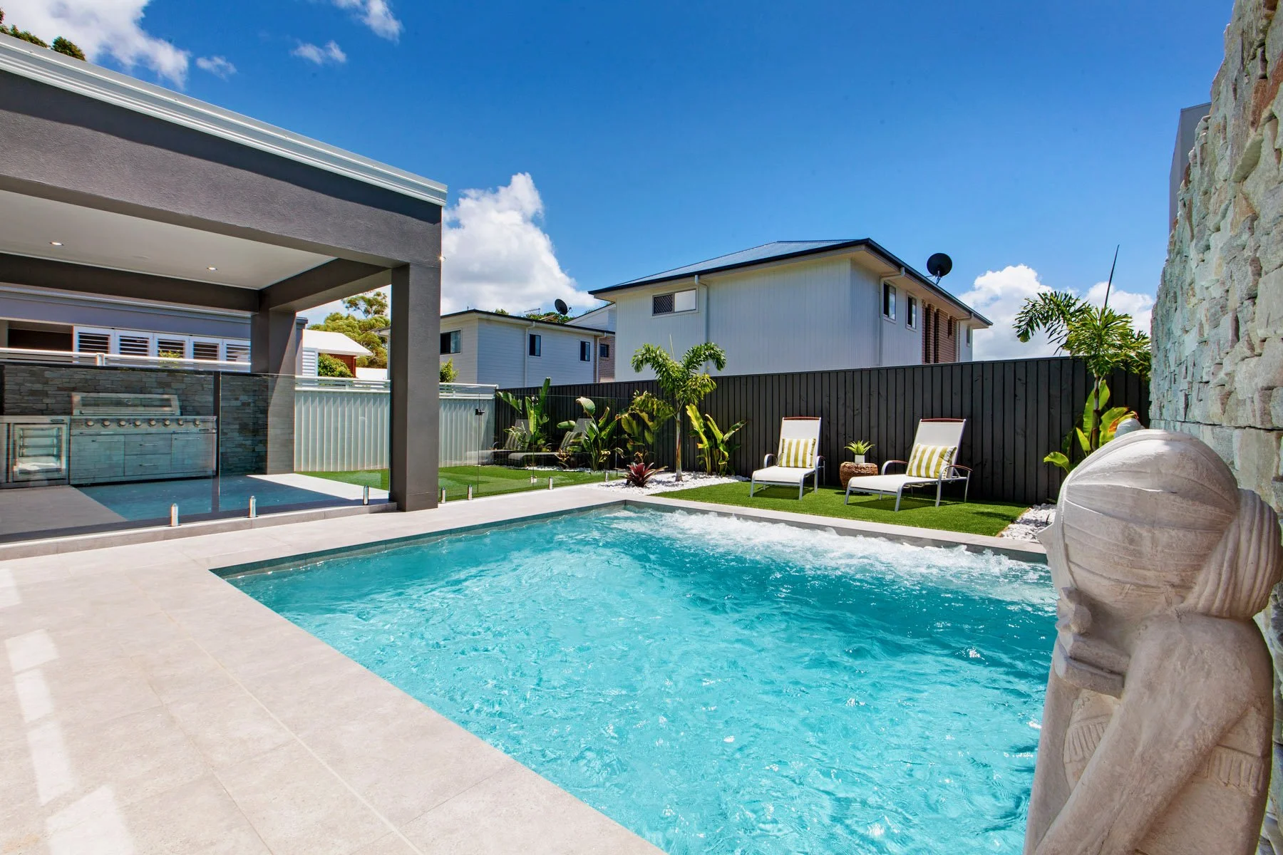A backyard with a swimming pool, two lounge chairs with striped cushions, a decorative water fountain, tropical plants, a black fence, modern houses, and a bright blue sky with clouds.