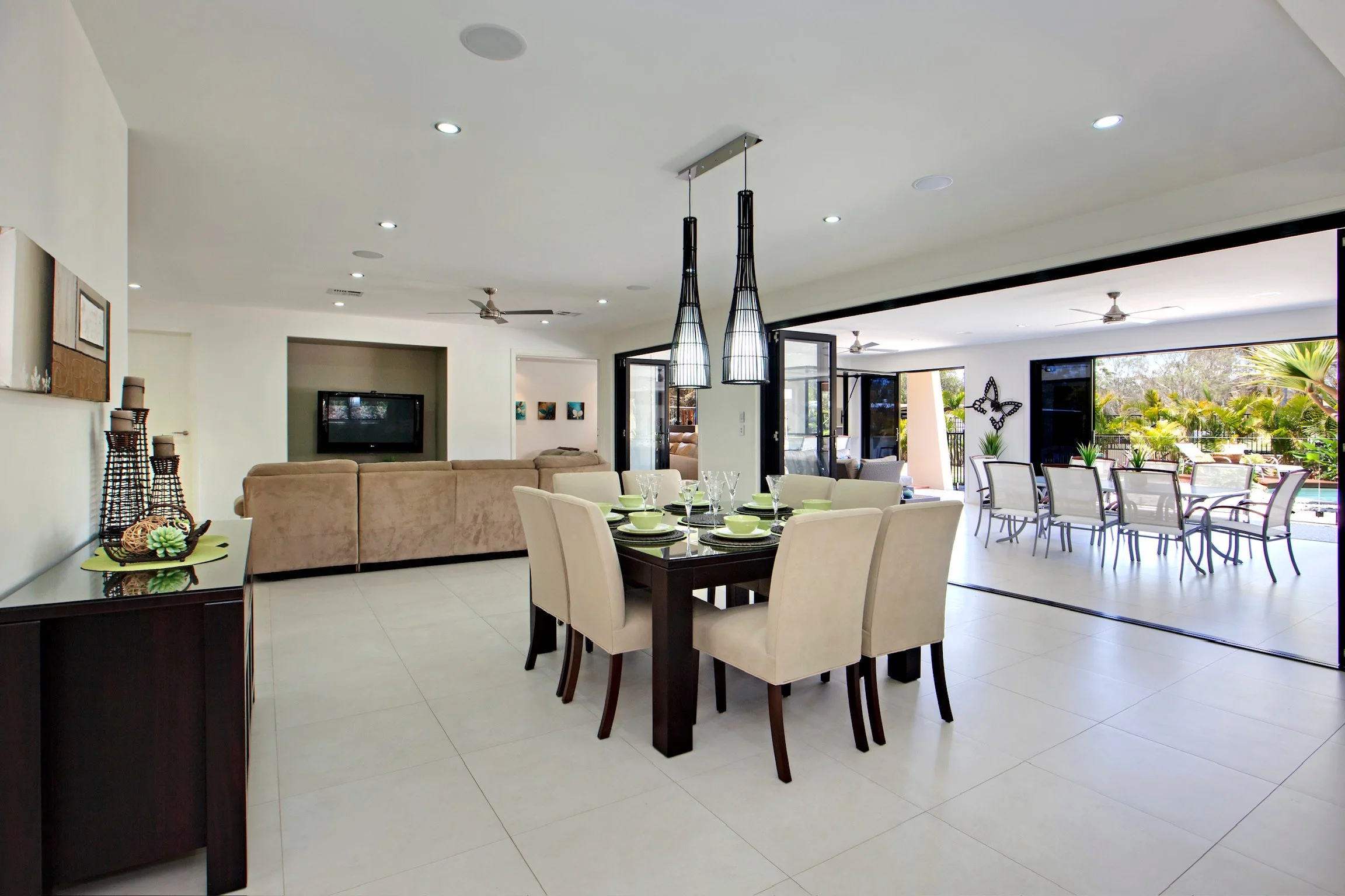 Open-plan living and dining room with sliding glass doors, white walls, tiled flooring, and modern furnishings, with outdoor patio and greenery visible.