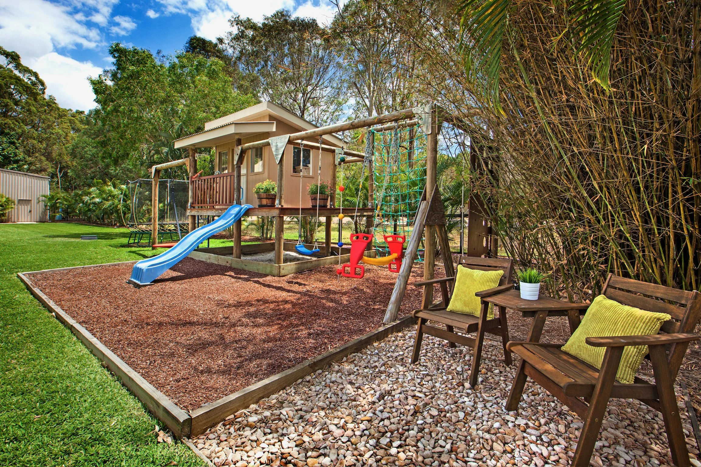 A backyard playground area with a small playhouse, a slide, a swing set, and a sandbox edged with wooden planks. There are two wooden chairs with green cushions and potted plants in the foreground. The area is surrounded by greenery, including tall t