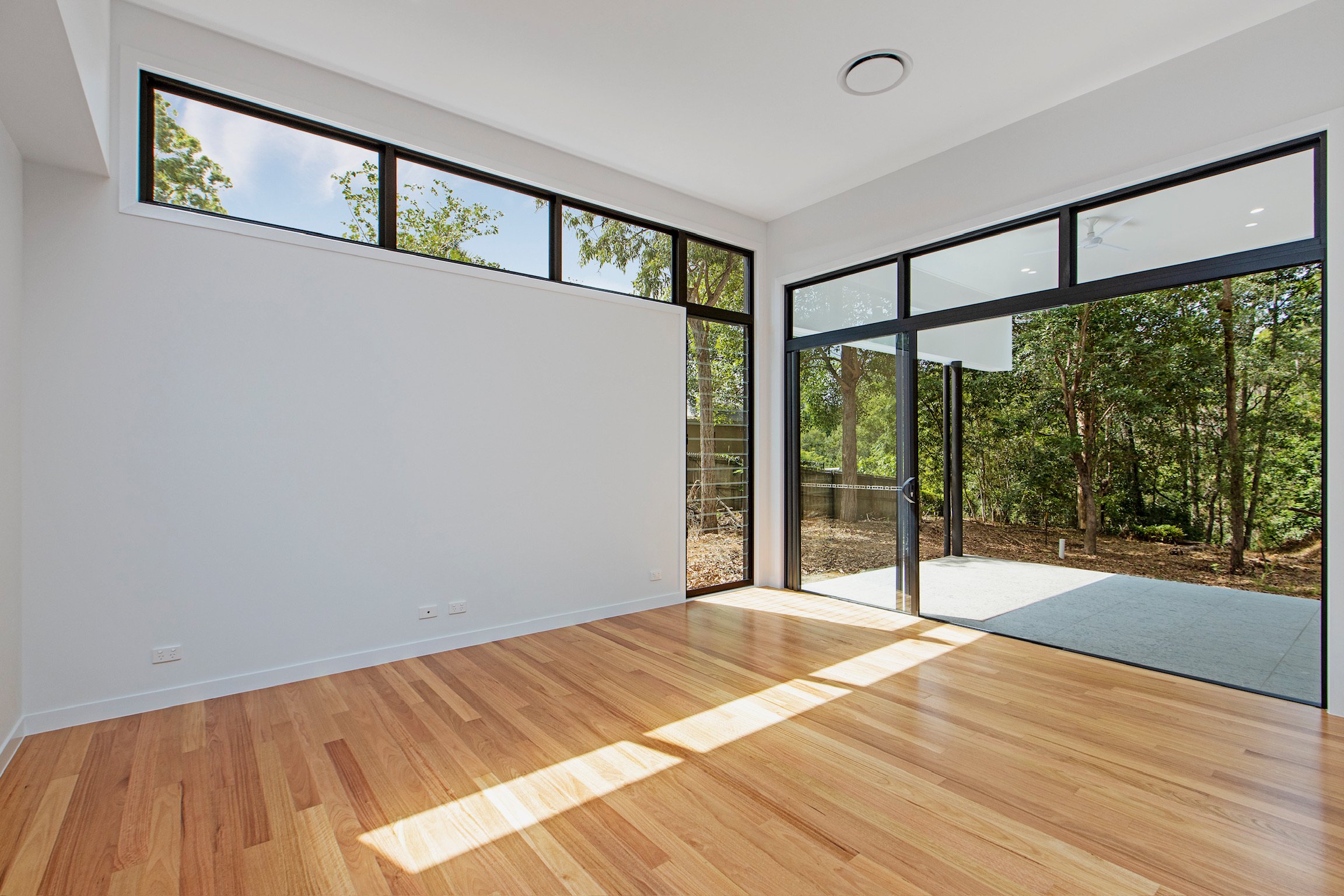 Empty modern living room with large floor-to-ceiling windows, wooden flooring, white walls, and view of trees outside.